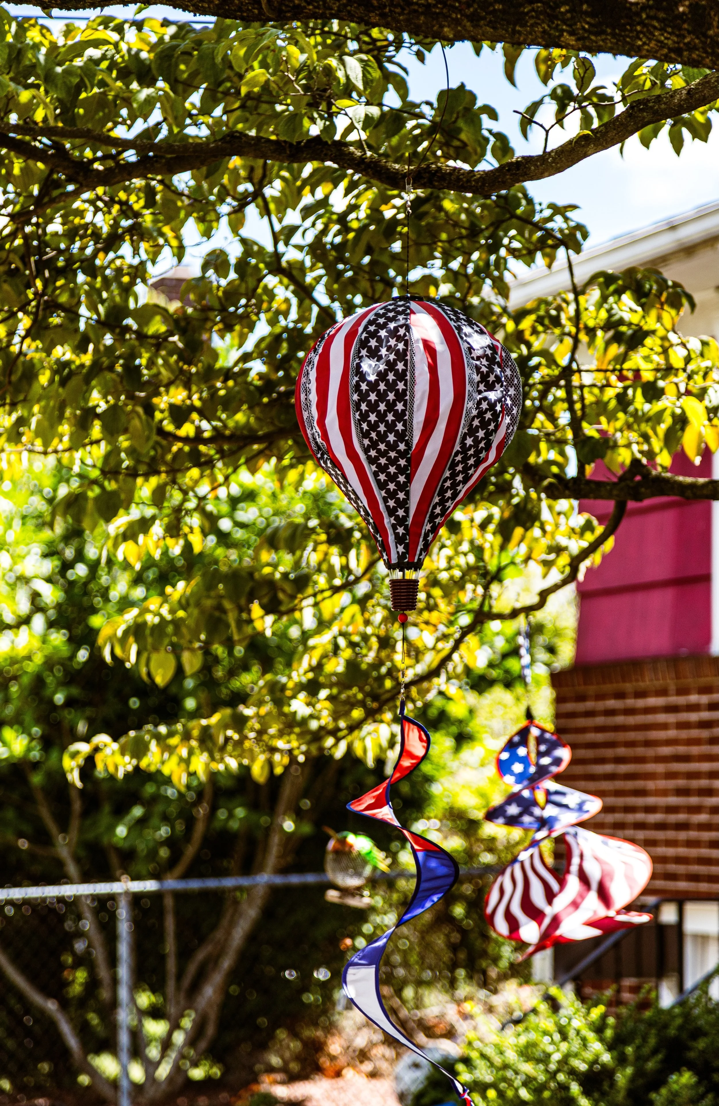 Colorful patriotic outdoor decorations, including a hot air balloon-shaped lantern and pinwheels with American flag designs, hanging from a tree branch on a sunny day.