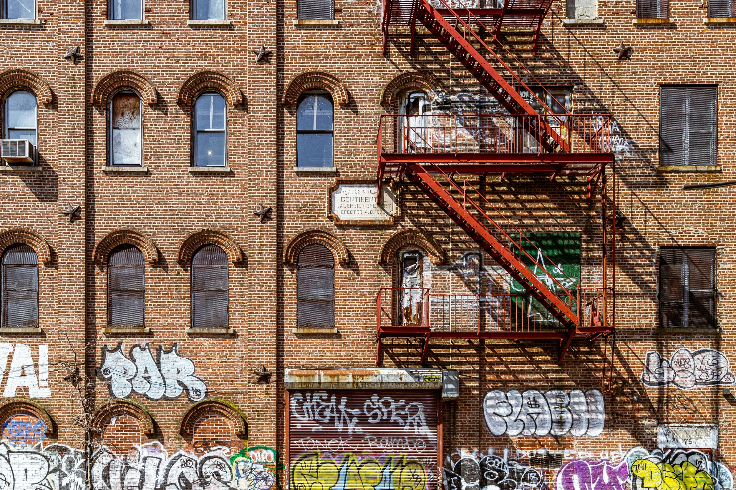 Brick building with multiple boarded-up windows, graffiti on the wall, and a red fire escape staircase.