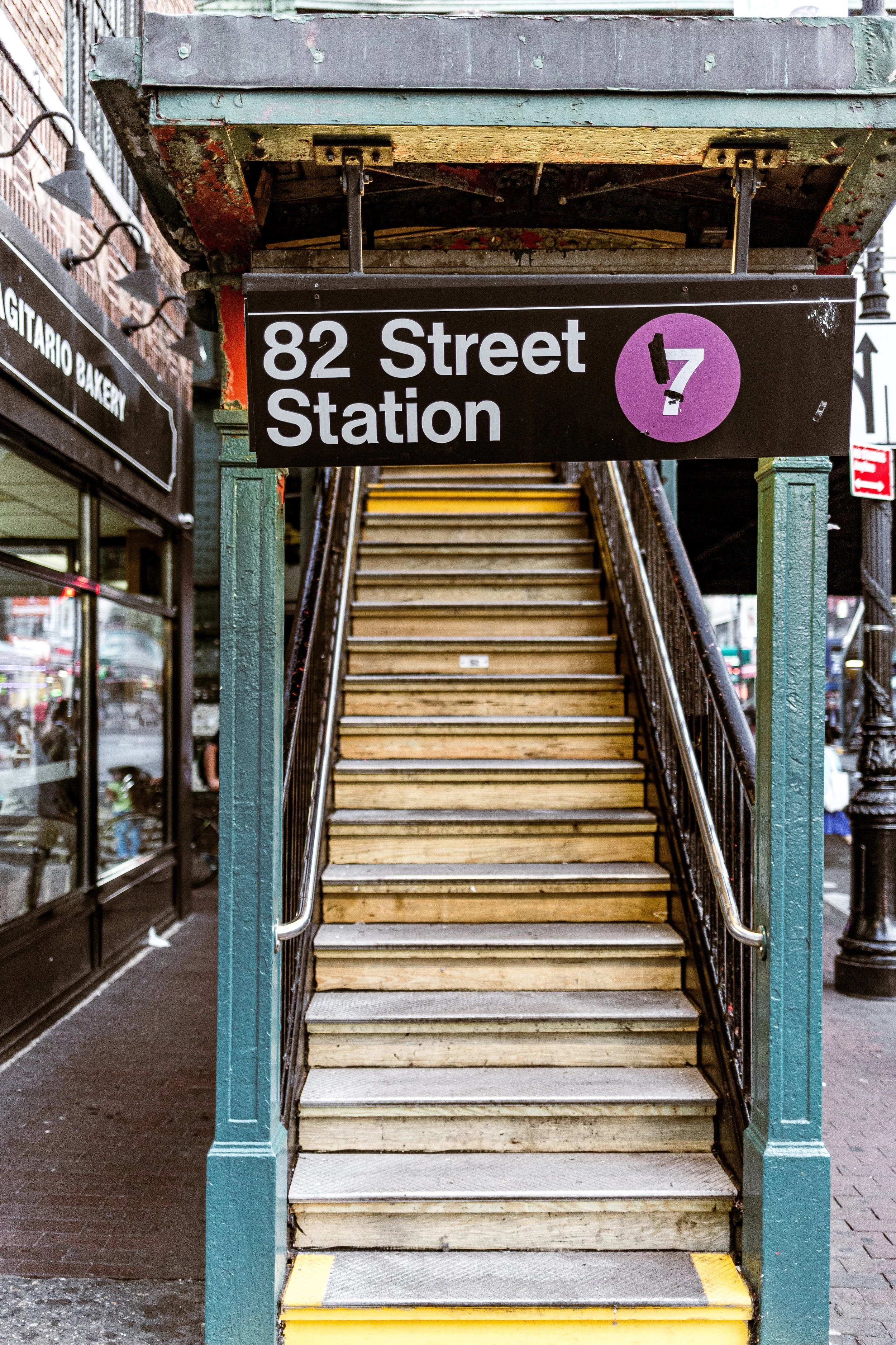 Outdoor staircase leading to 82 Street Station with a black sign above, featuring bold white text and a purple circle with a black symbol.