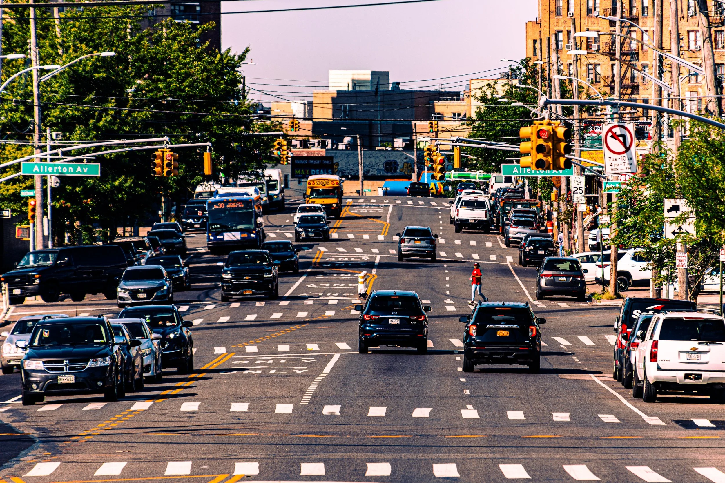 A busy city intersection with cars, trucks, and pedestrians on Allerton Avenue, traffic lights, and buildings along the street.