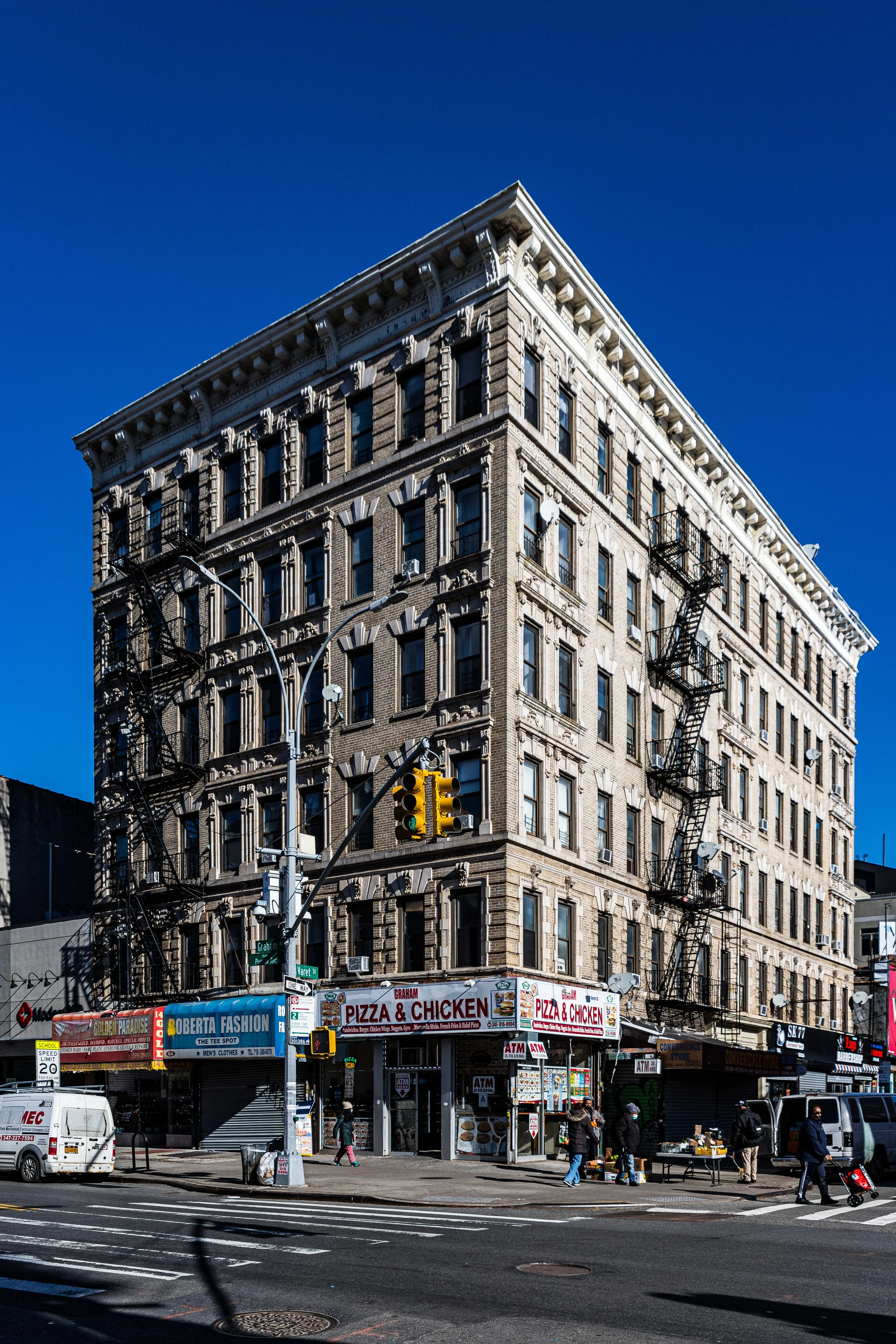 A multi-story beige brick building with fire escapes on each floor, located on a city street corner. Ground level has signage for pizza and chicken, along with ATMs and small shops. Pedestrians are walking along the sidewalk, and a traffic light is visible in front of the building.