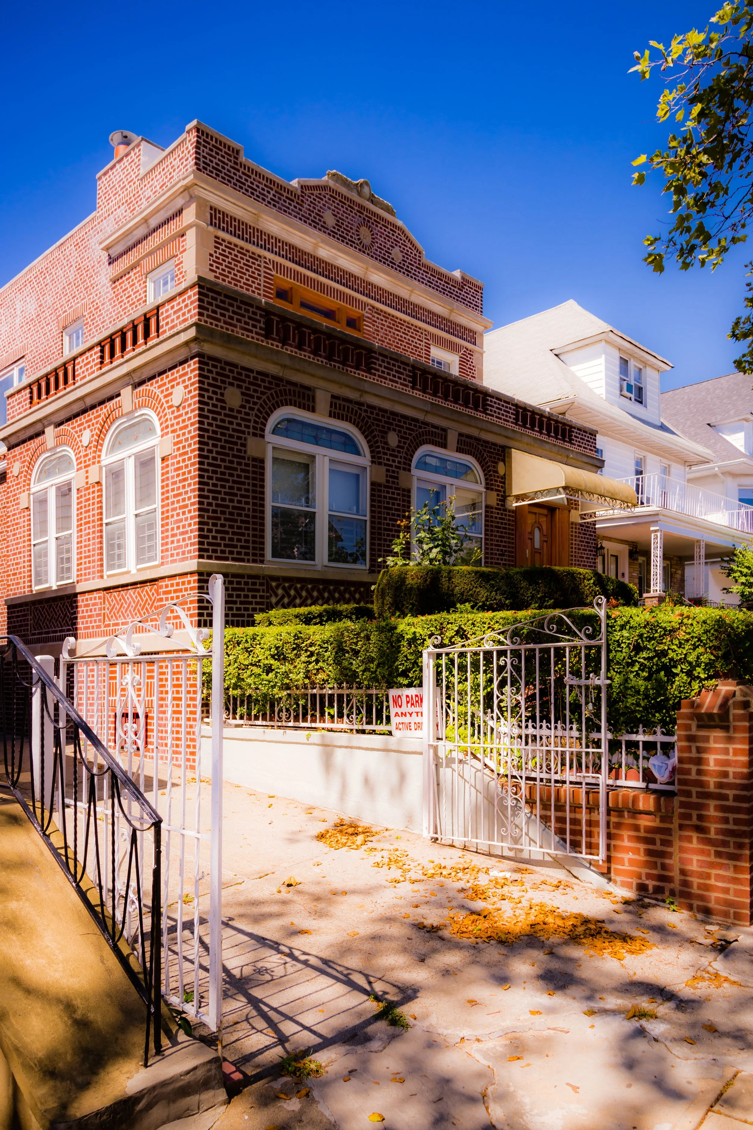 A red brick multi-story house with arched windows and a small garden, surrounded by a white iron gate and fence, under a clear blue sky.