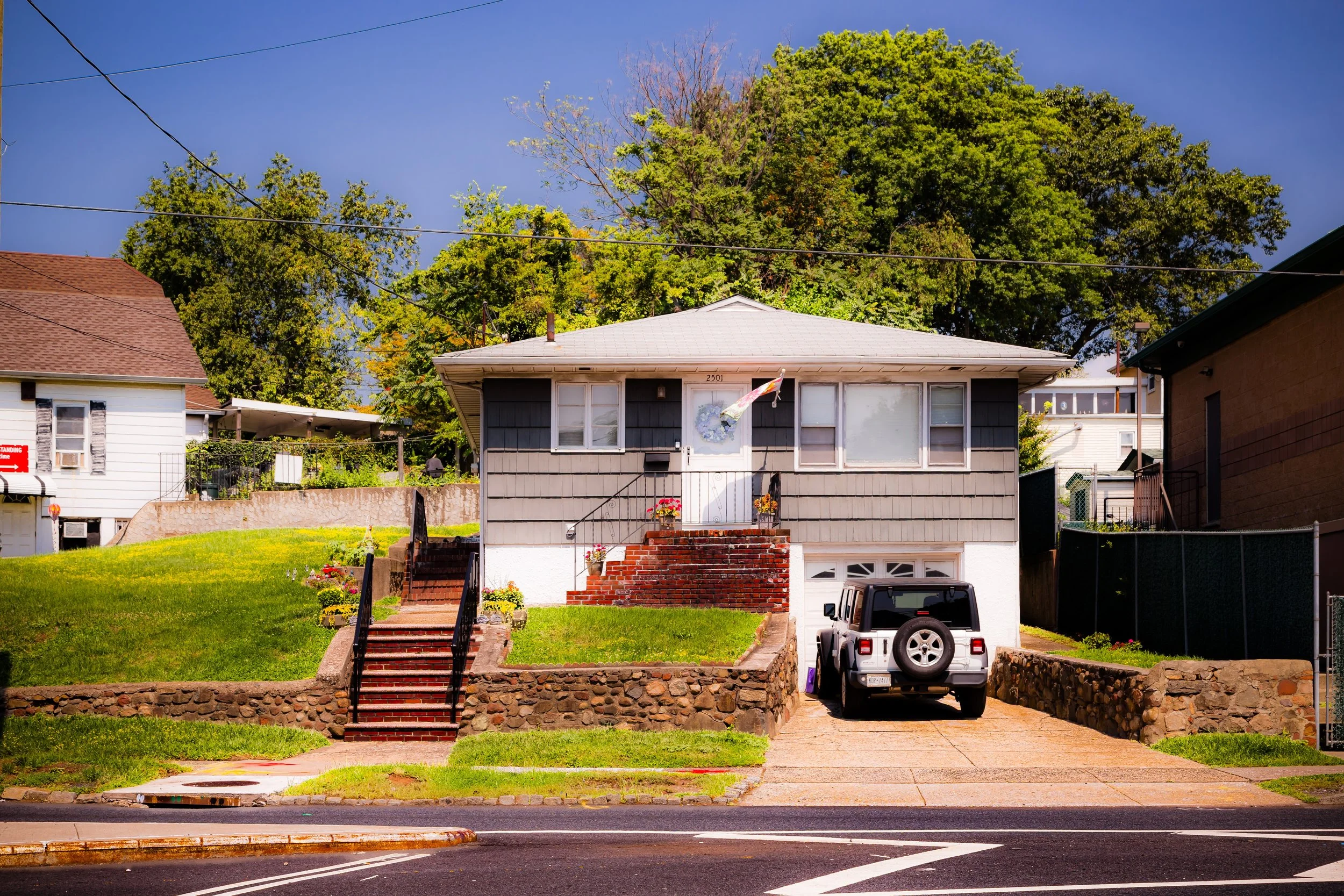 A two-story house with gray siding, front stairs, a garage, and a white door, set on a grassy hill with trees in the background.