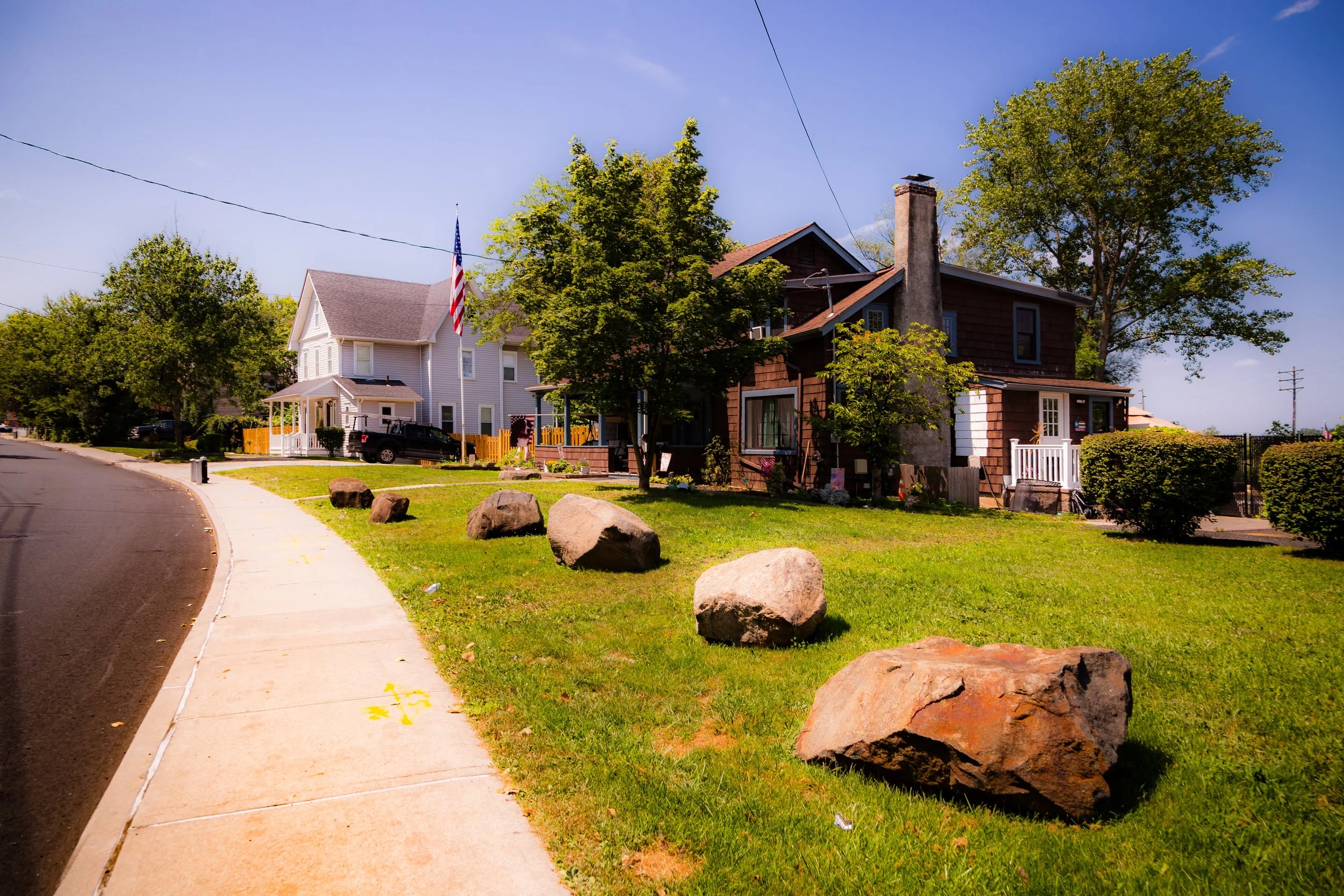 Residential street corner with houses, trees, a sidewalk, and large rocks in the grass, under a clear blue sky.