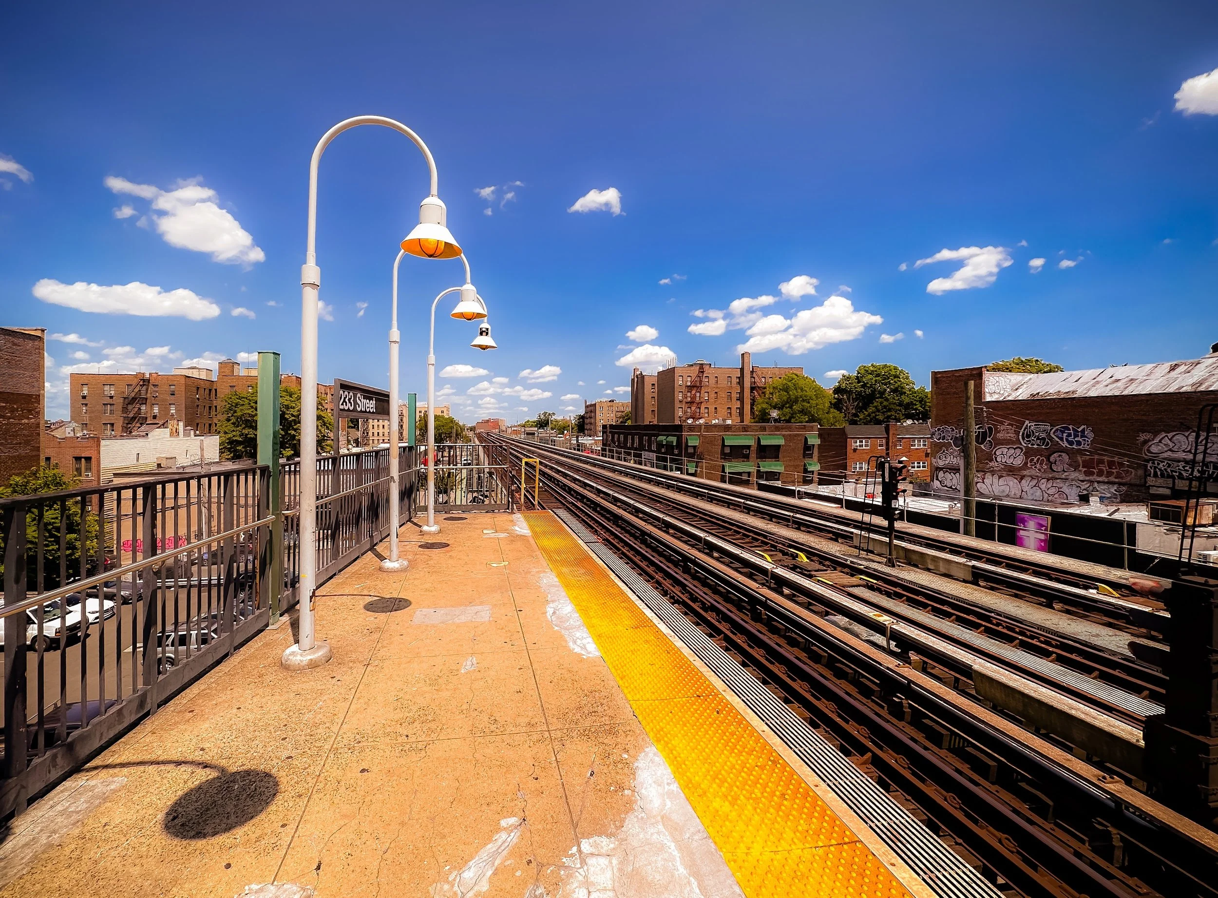 View of a train station platform with yellow safety strip, white lamps, and railway tracks under a blue sky with scattered clouds.