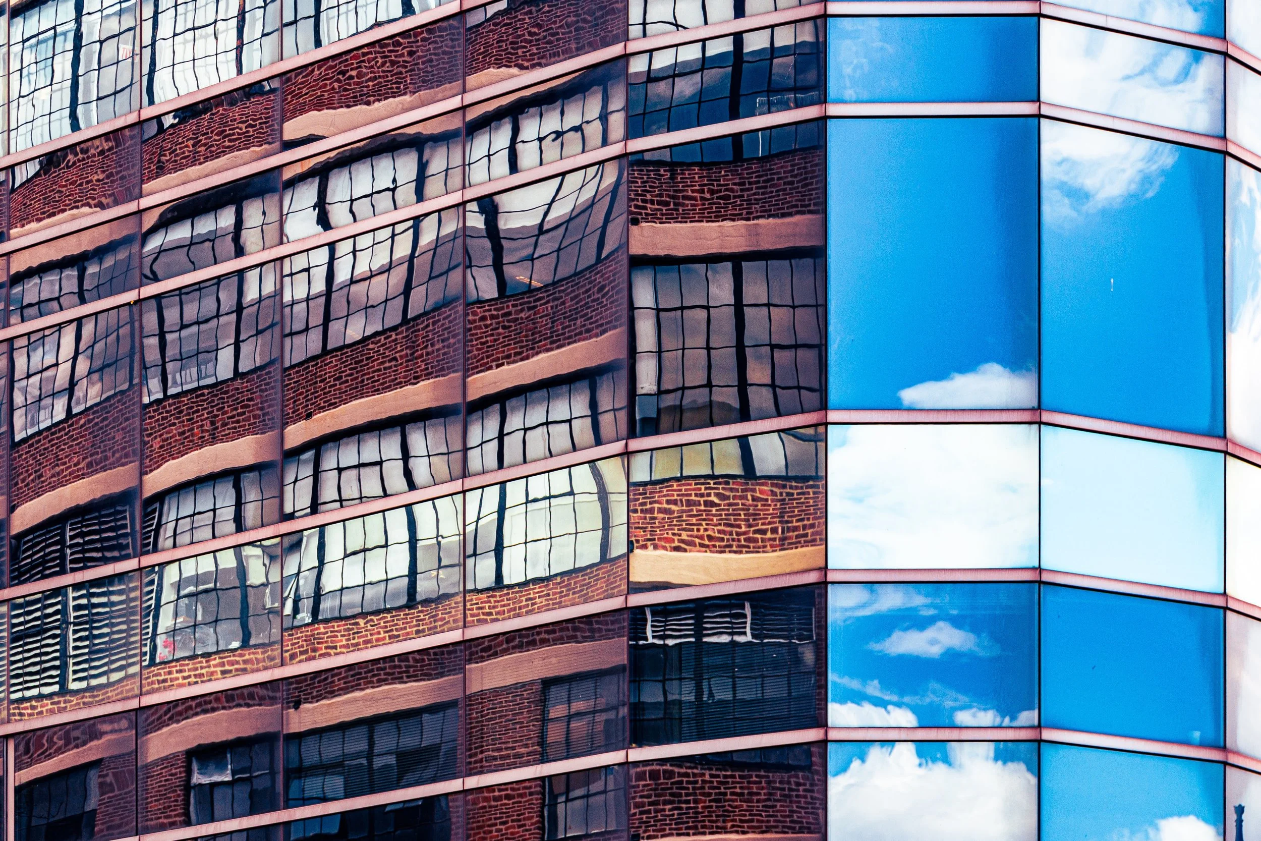 Reflections of buildings and sky on a modern glass building facade with blue, cloudy sky and brick structures.
