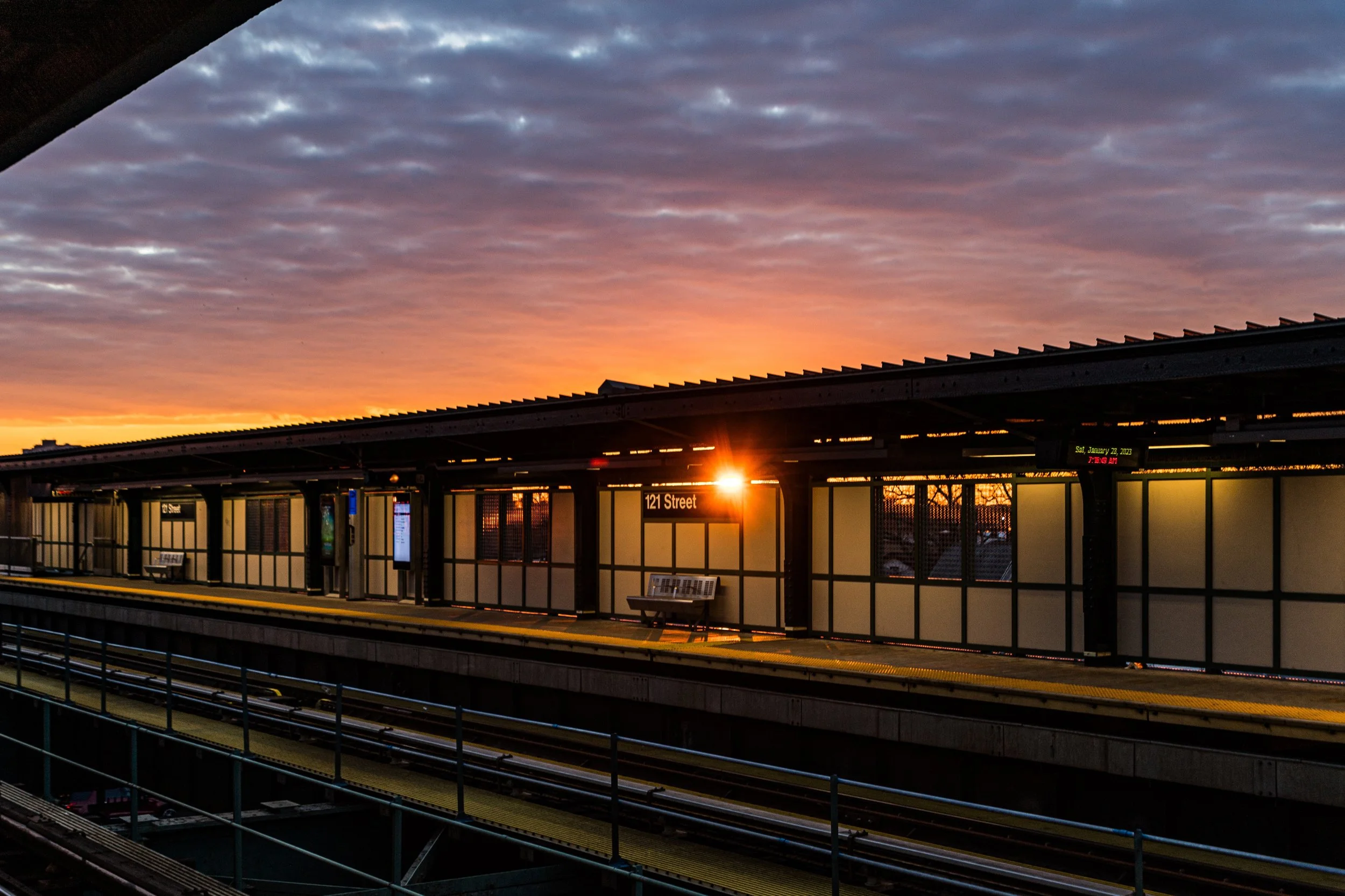 Subway station platform at sunset, with a sign indicating '121 Street,' benches, digital displays, and train tracks in the foreground.