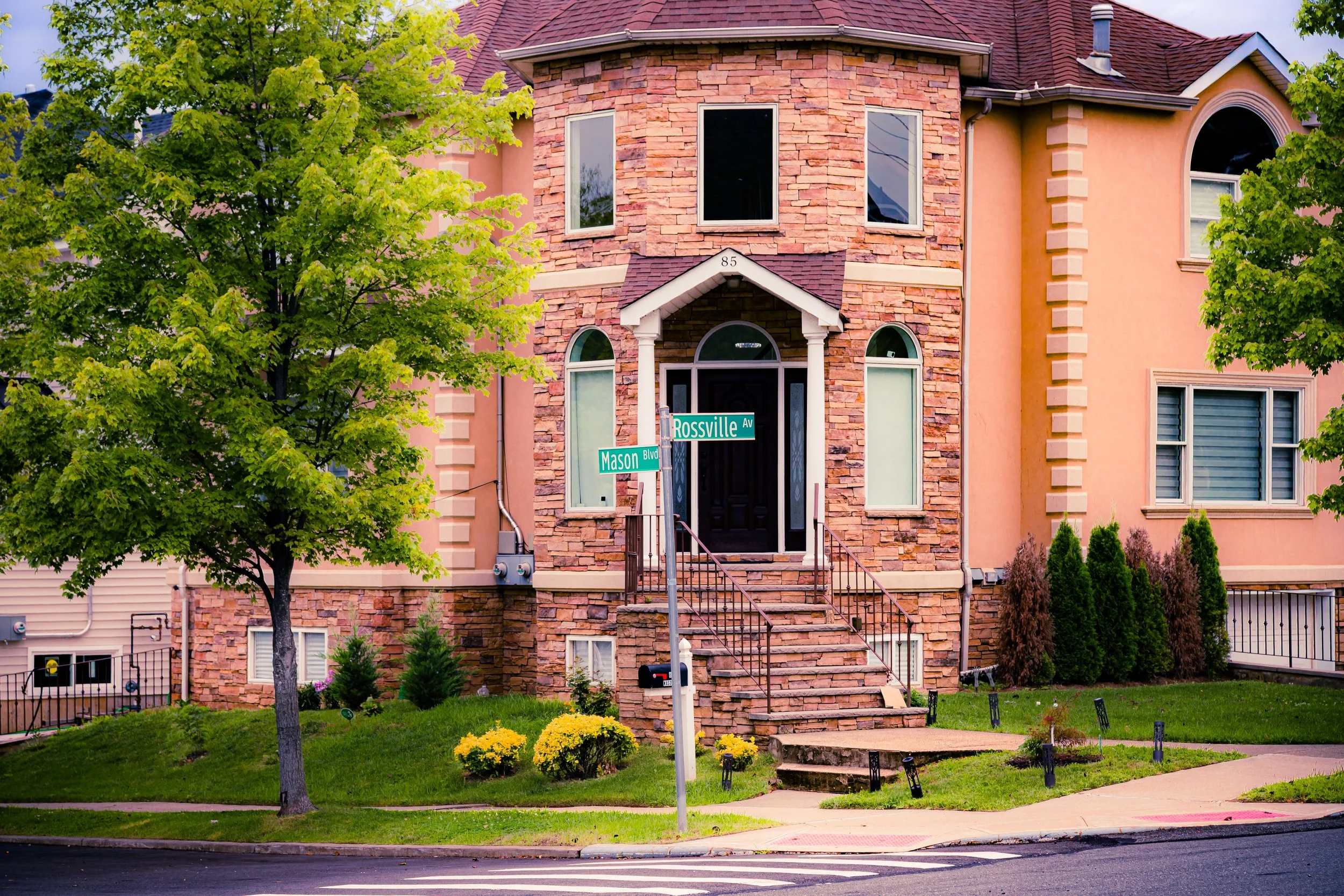 Colorful two-story house at corner of Mason Blvd and Rossville Ave, with brick and pink walls, stairs leading to front door, green trees, grass, and street signs.