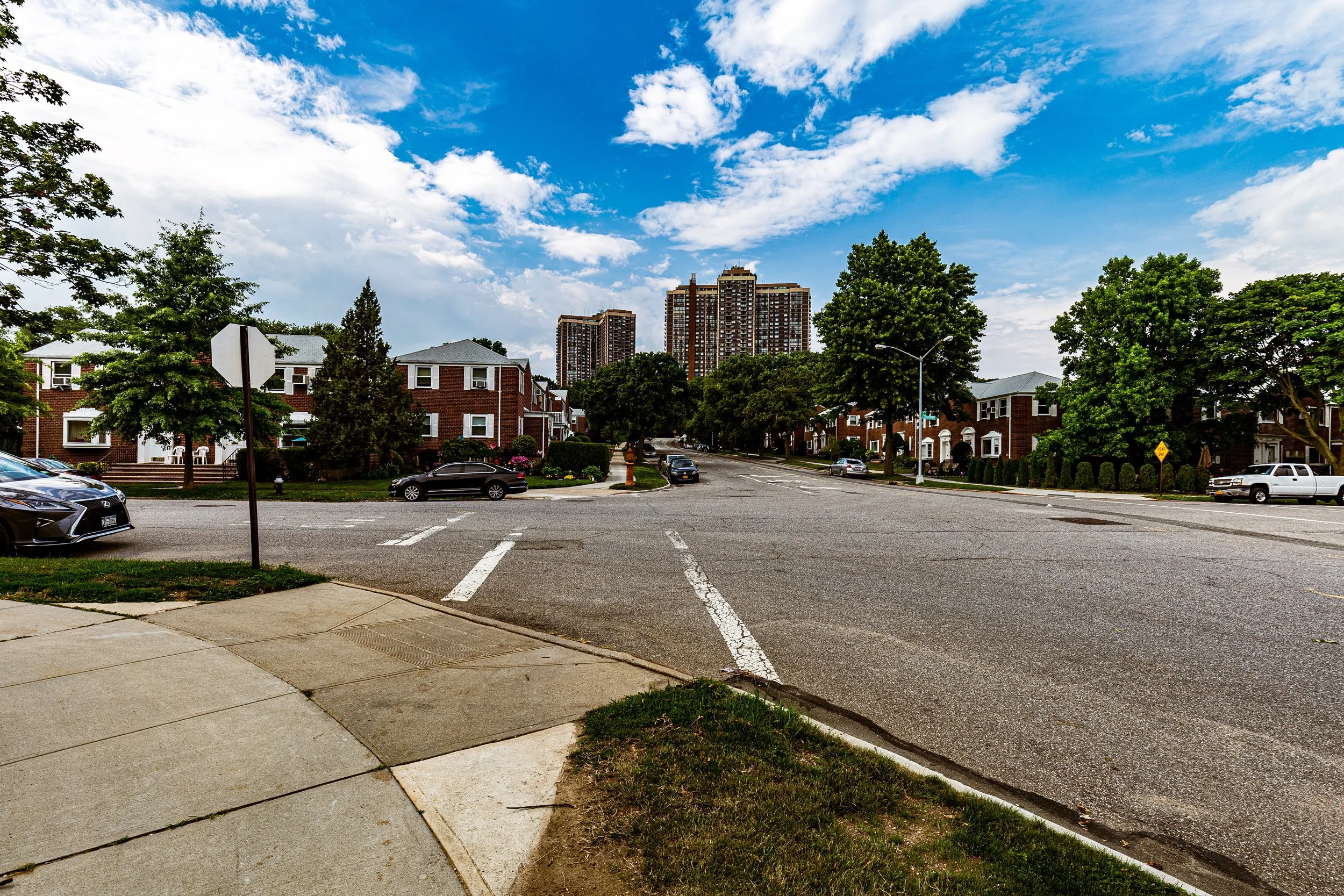 A street intersection in a suburban neighborhood with residential buildings and taller apartment complexes in the background, green trees lining the street, parked cars, and a partly cloudy blue sky.