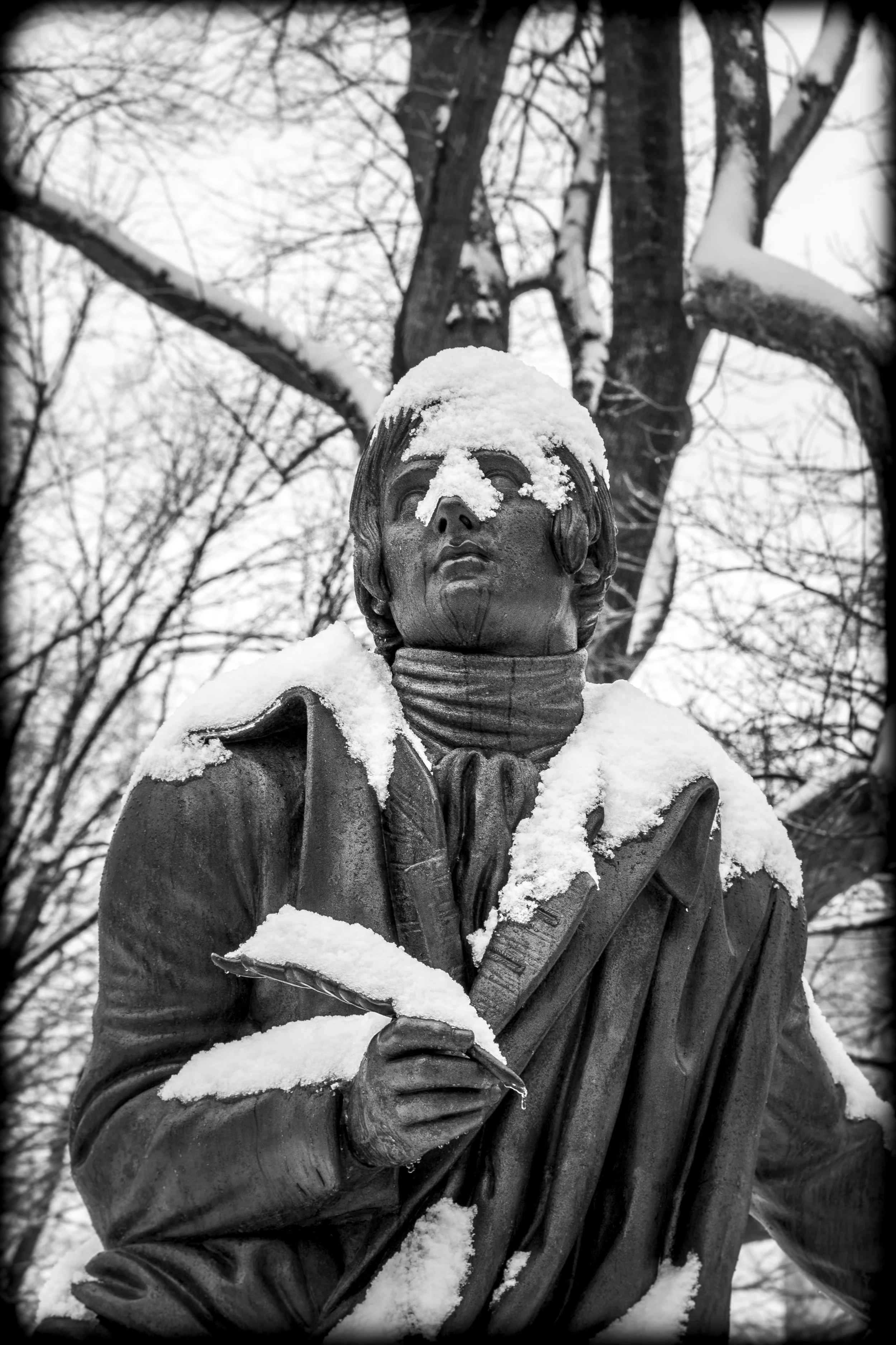 Snow-covered statue of a man holding a paper, with snow on his face and shoulders, and bare tree branches in the background.