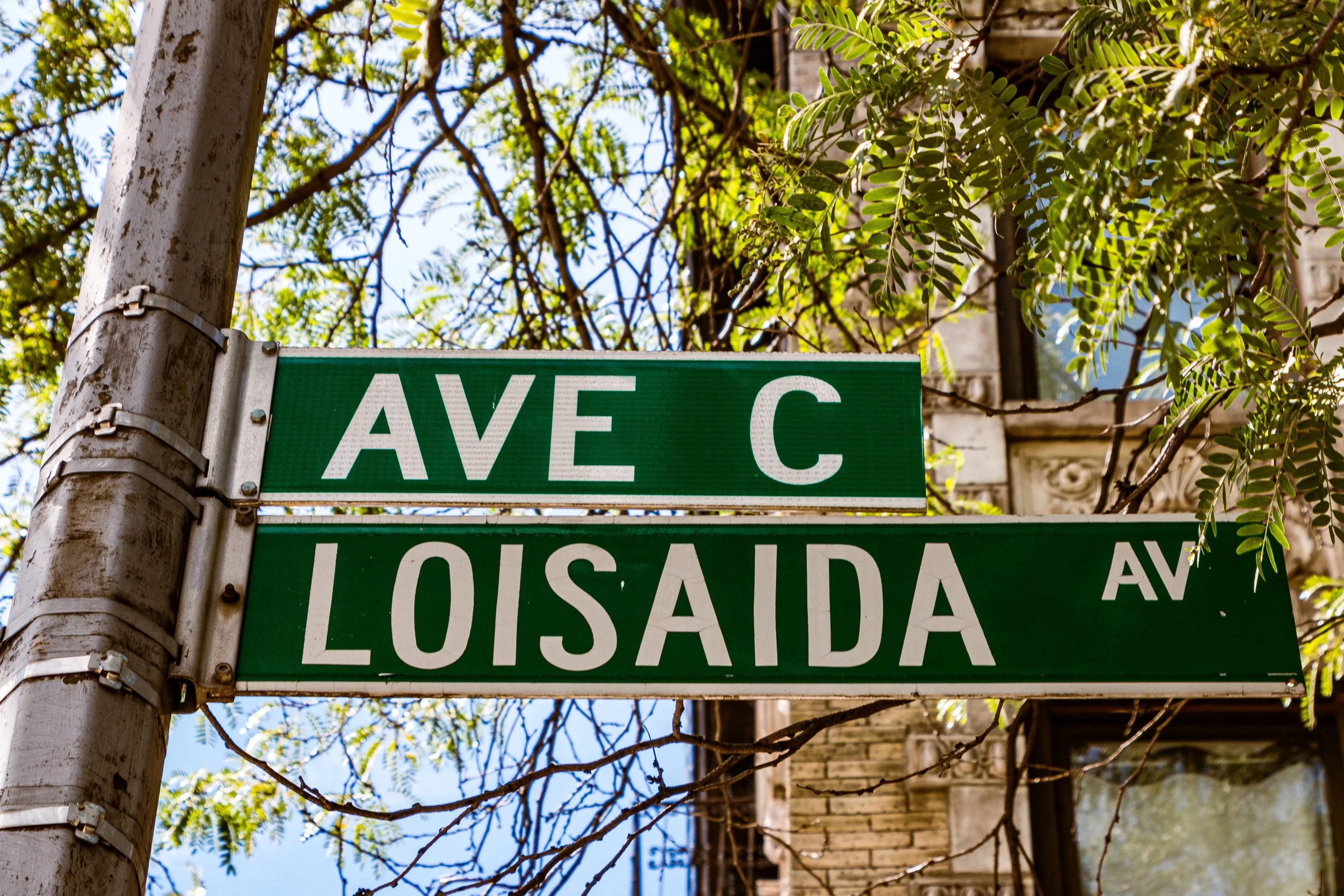 Street signs reading 'AVÉ C' and 'LOISADA AV' attached to a metal pole, with trees and a building in the background.
