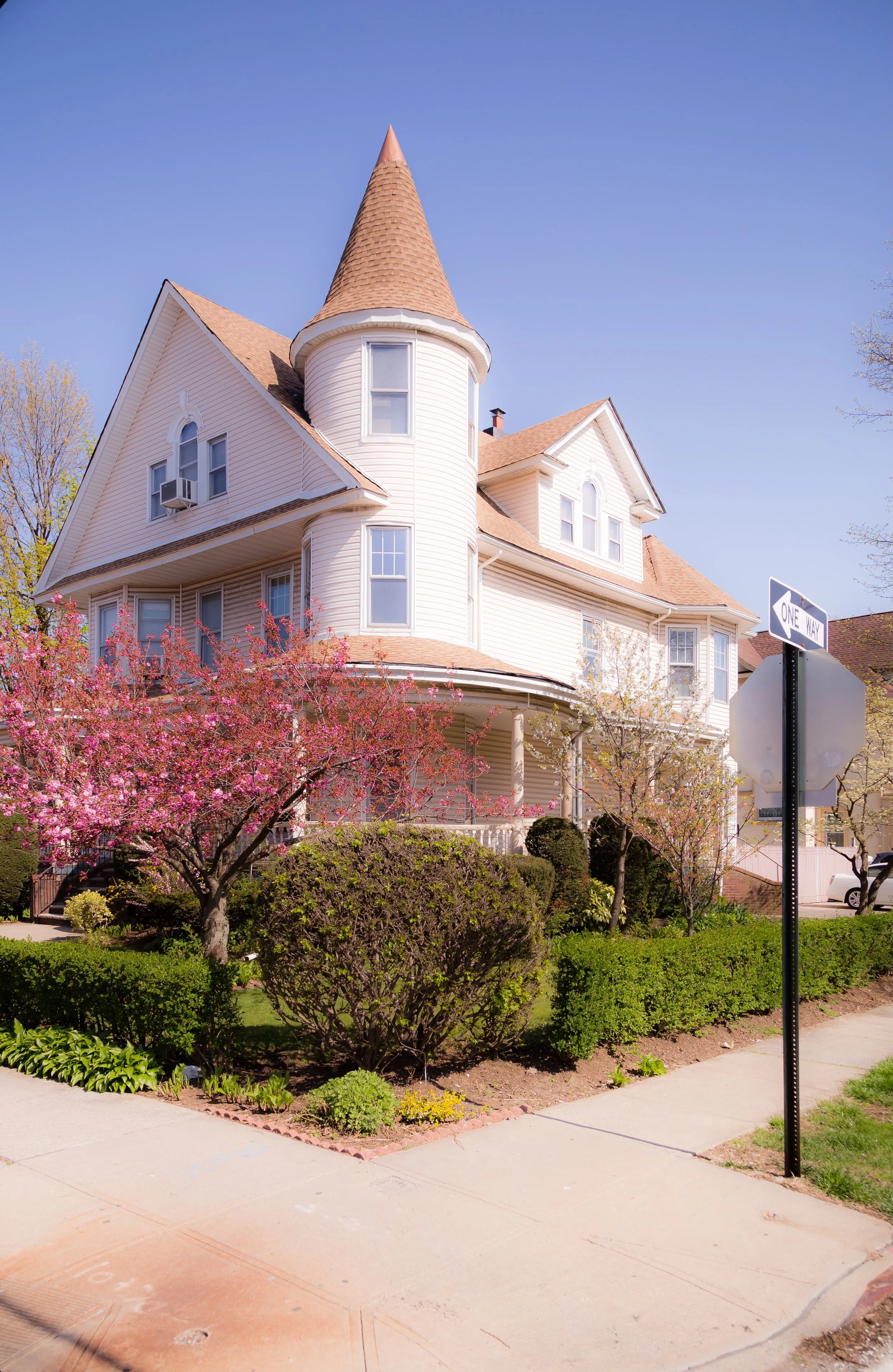 A large Victorian-style house painted white with a conical tower, surrounded by blooming pink cherry blossom trees and green shrubs on a sunny day.