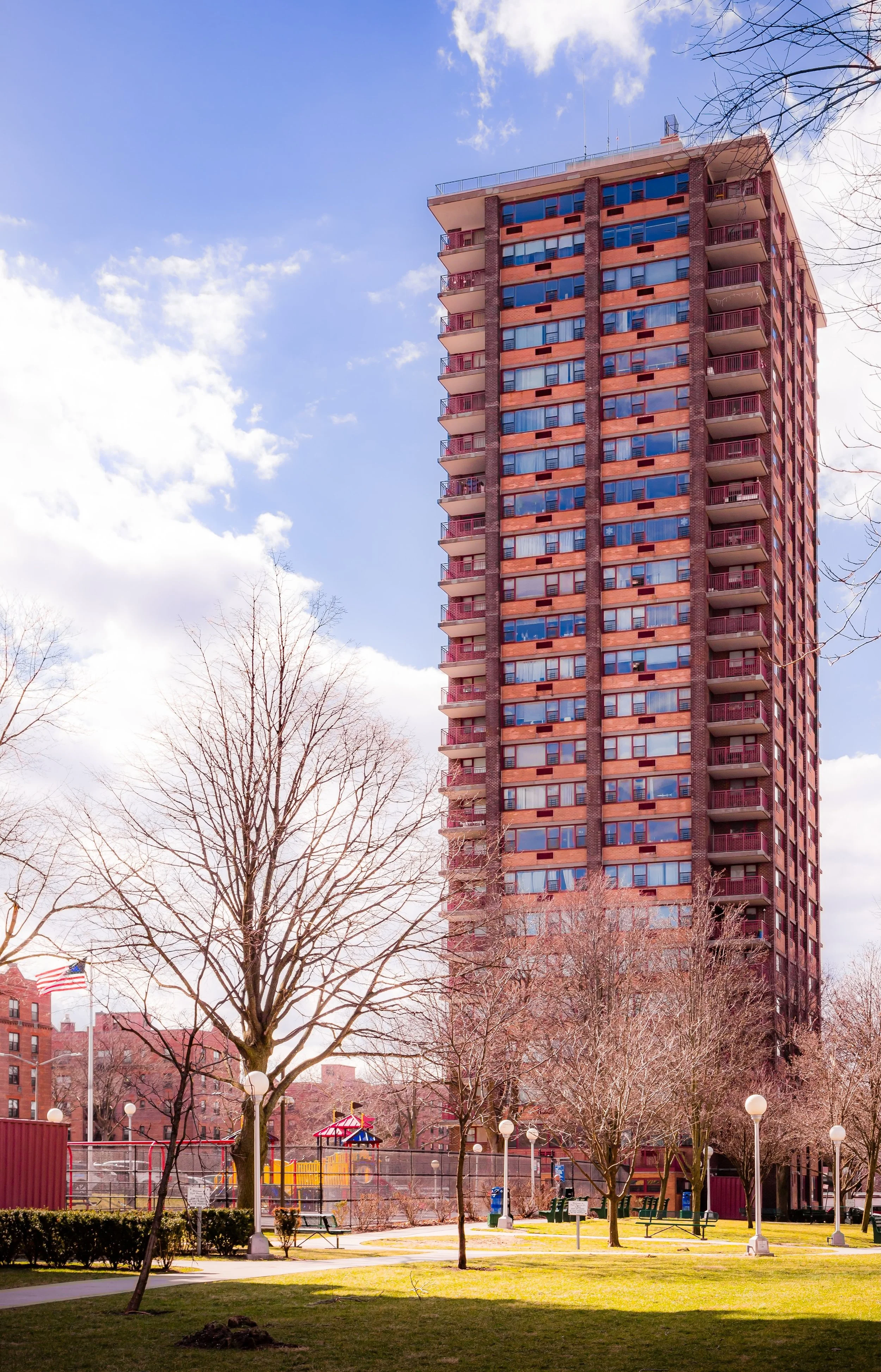A tall apartment building with a brick facade, balconies on each floor, and blue-tinted windows. There are leafless trees and a park with a playground, benches, lamps, and a flag in the foreground. The sky is partly cloudy.