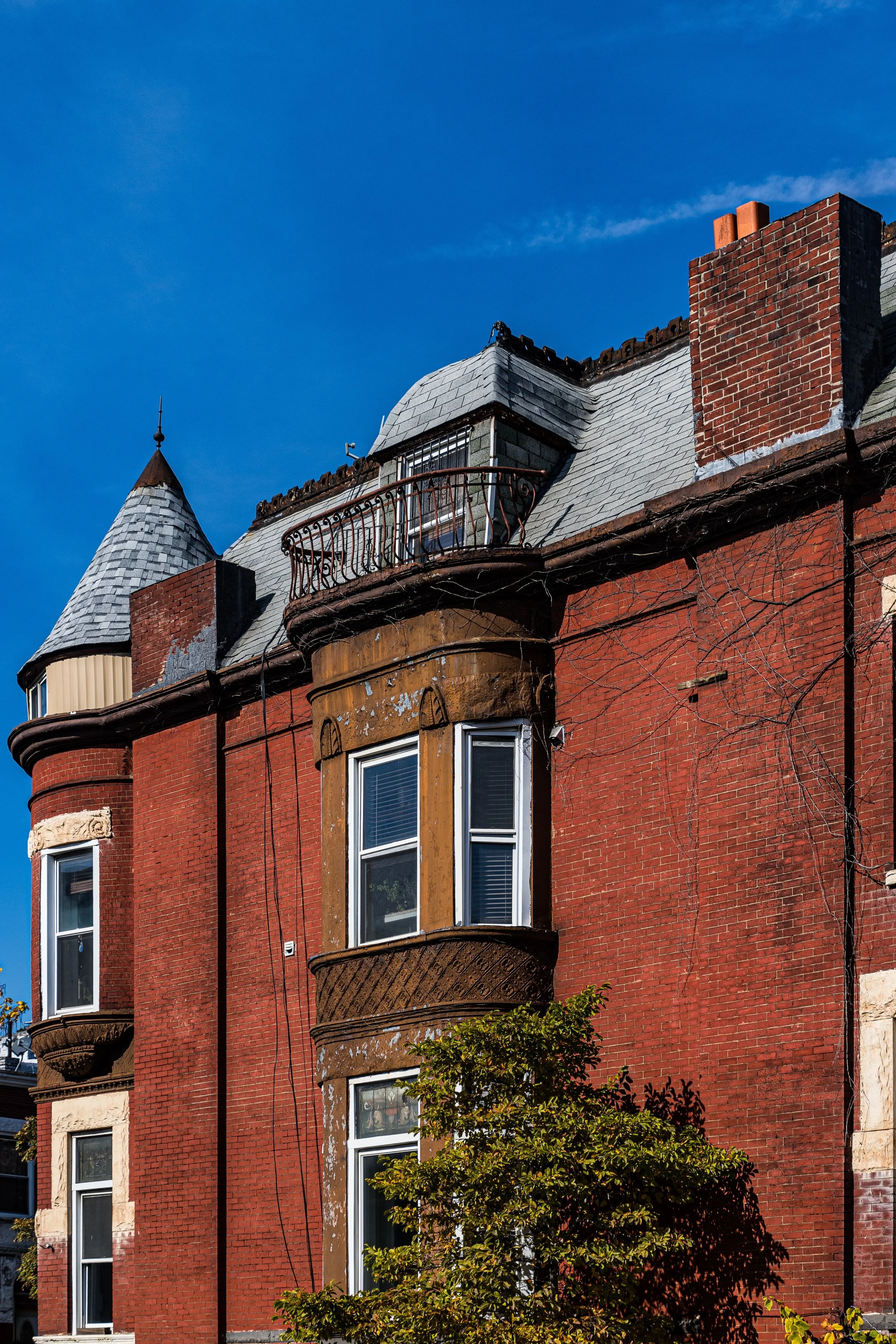 Close-up of a red brick Victorian-style building with turret, dormer windows, a small balcony, and leafless vines against a blue sky.