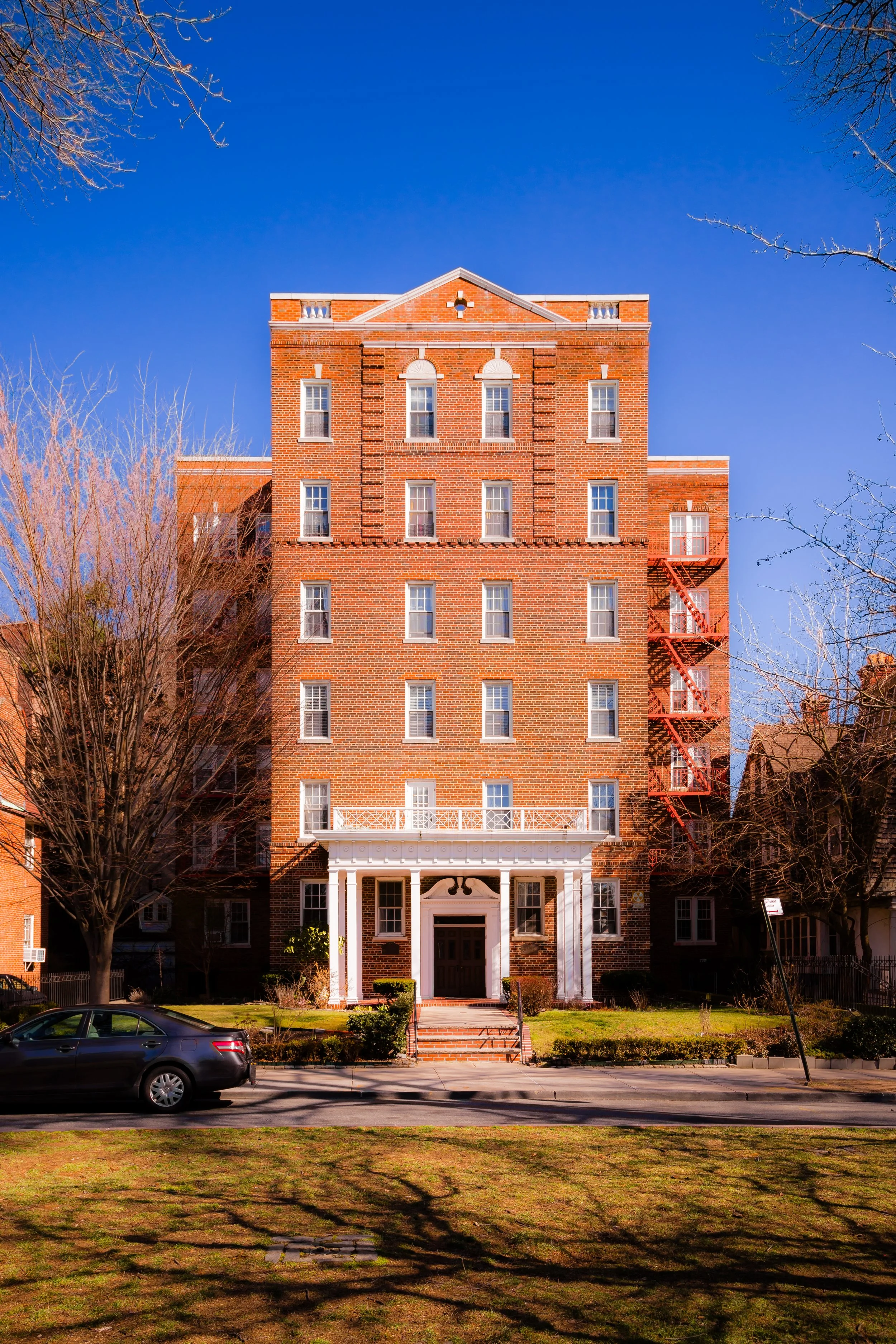 A multi-story brick building with a white portico entrance and fire escape stairs on the right side, set against a bright blue sky with leafless trees in the foreground.