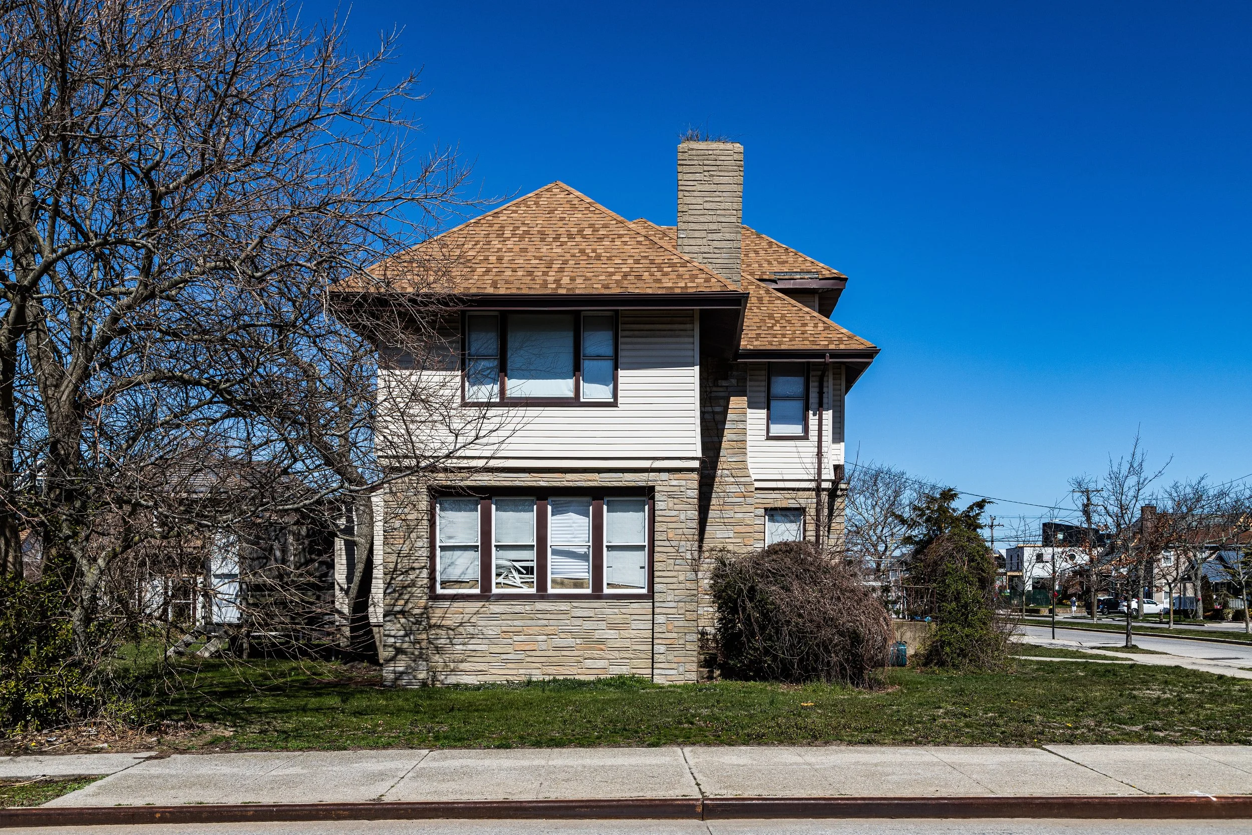 A two-story house with beige stone exterior, white siding, and brown shingle roof, situated on a lawn with leafless trees and bushes, under a clear blue sky.