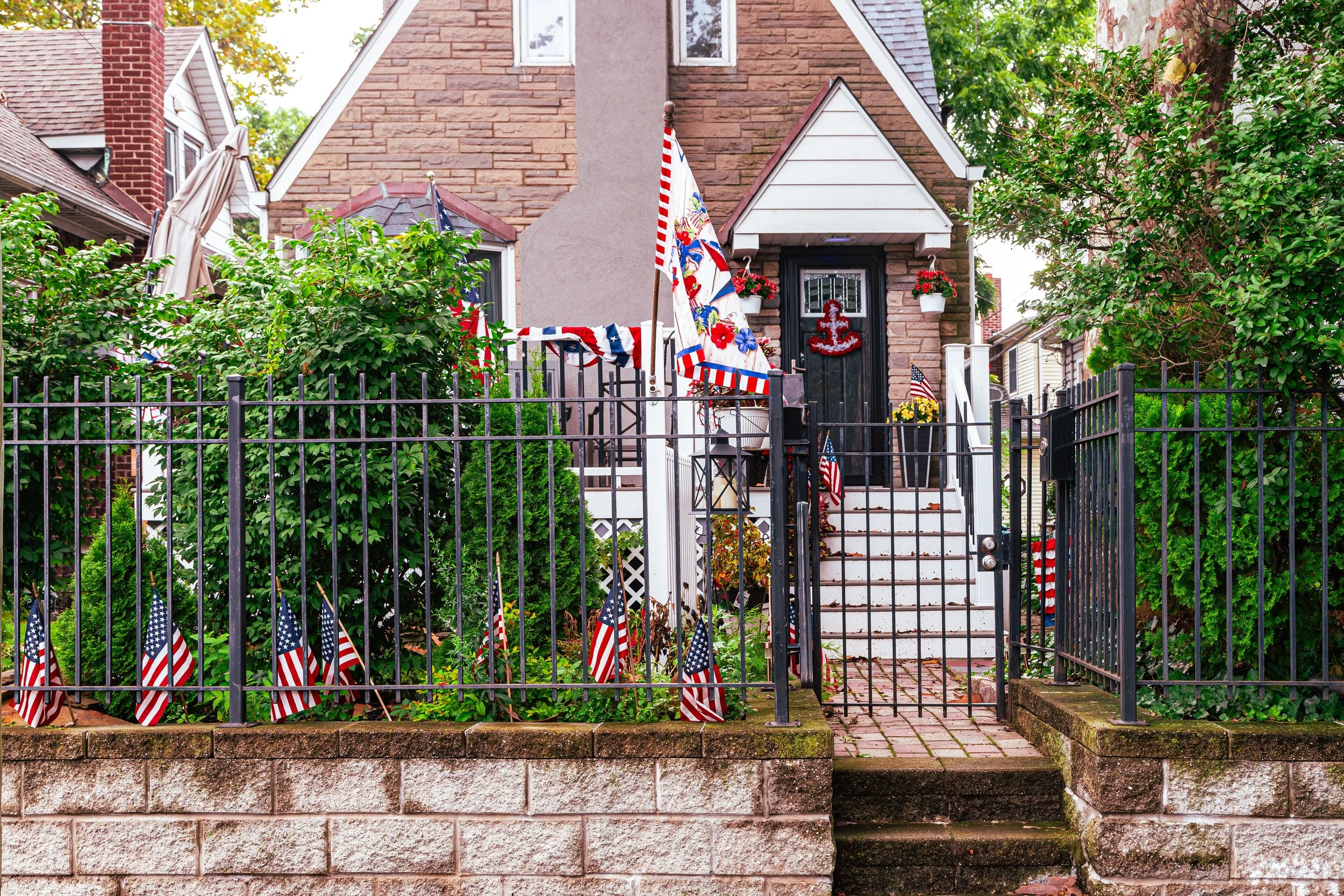 A house decorated with American flags, patriotic banners, and red, white, and blue decorations for a patriotic holiday, with a black front door and white staircase, surrounded by lush green bushes and trees.