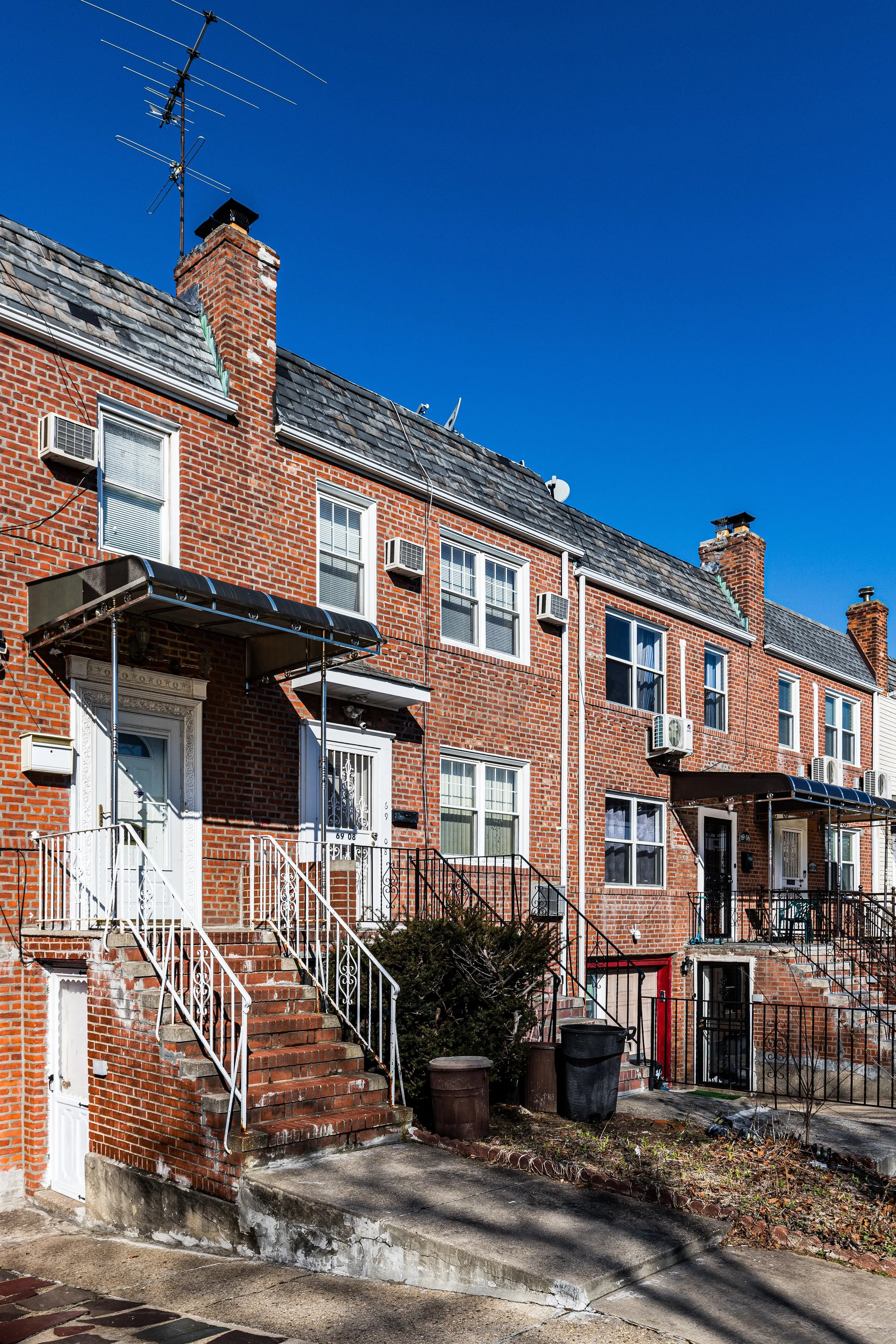 A row of brick townhouses with front stairs, small porches, and multiple windows, under a clear blue sky.