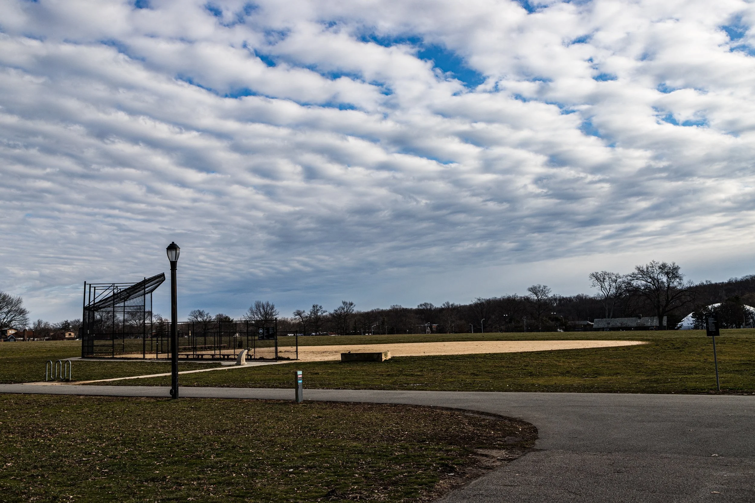 A park with a baseball field, a lamp post, a sidewalk, and trees in the background under a partly cloudy sky.