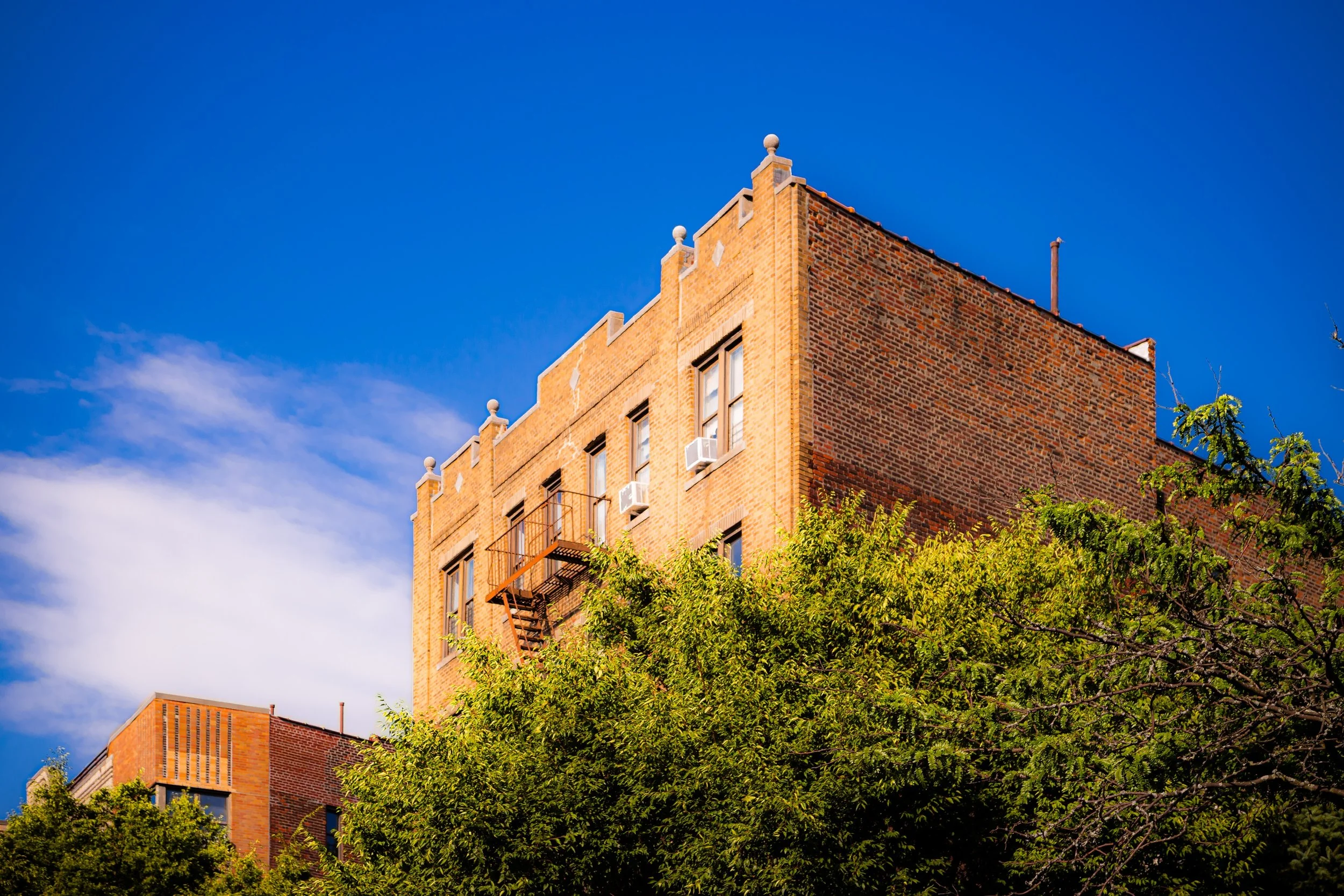 A brick building with multiple windows and fire escape stairs, partially obscured by green trees against a bright blue sky with some clouds.