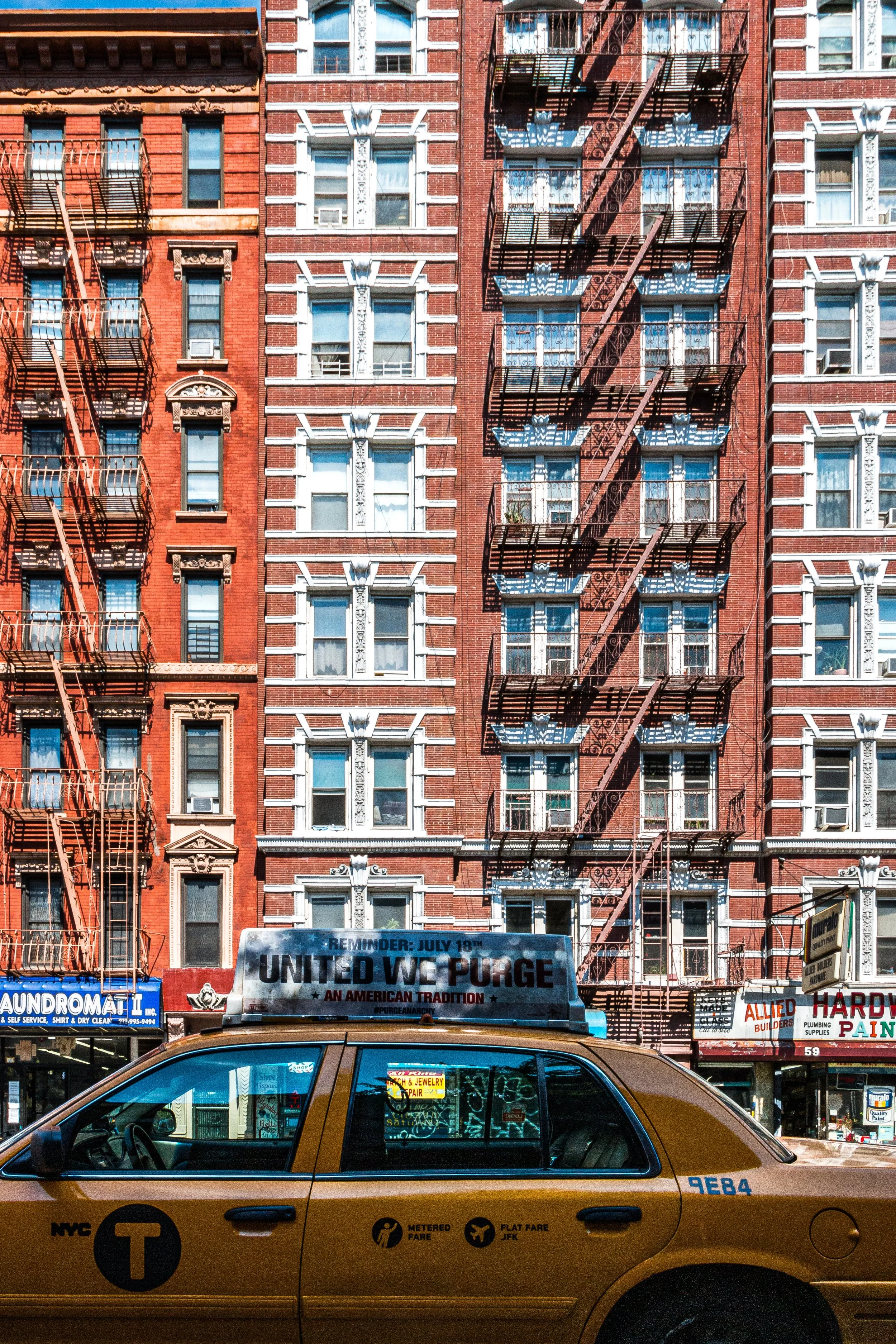 Yellow NYC taxi cab parked in front of a brick apartment building. A sign on the car's roof reads, 'REMINDER: JULY 18th, UNITED WE PURGE, AN AMERICAN TRADITION.' The building features fire escape stairs and various shop signs at street level.