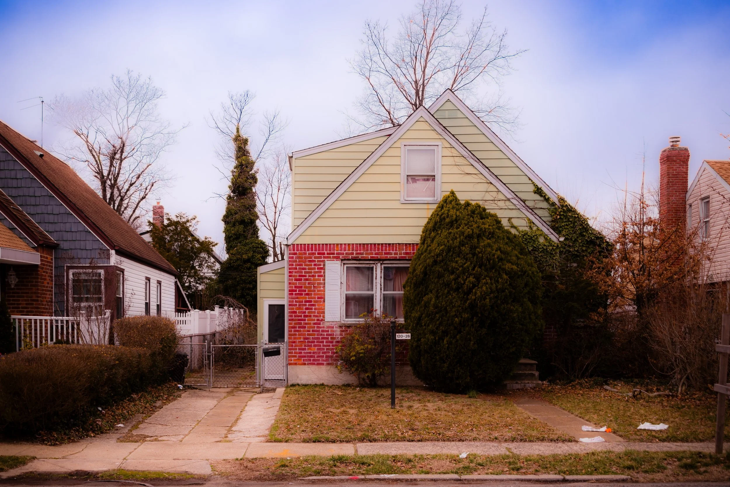 A two-story house with a brick and siding exterior, large bushes in front, and leafless trees in the background, under a pink and purple sky.