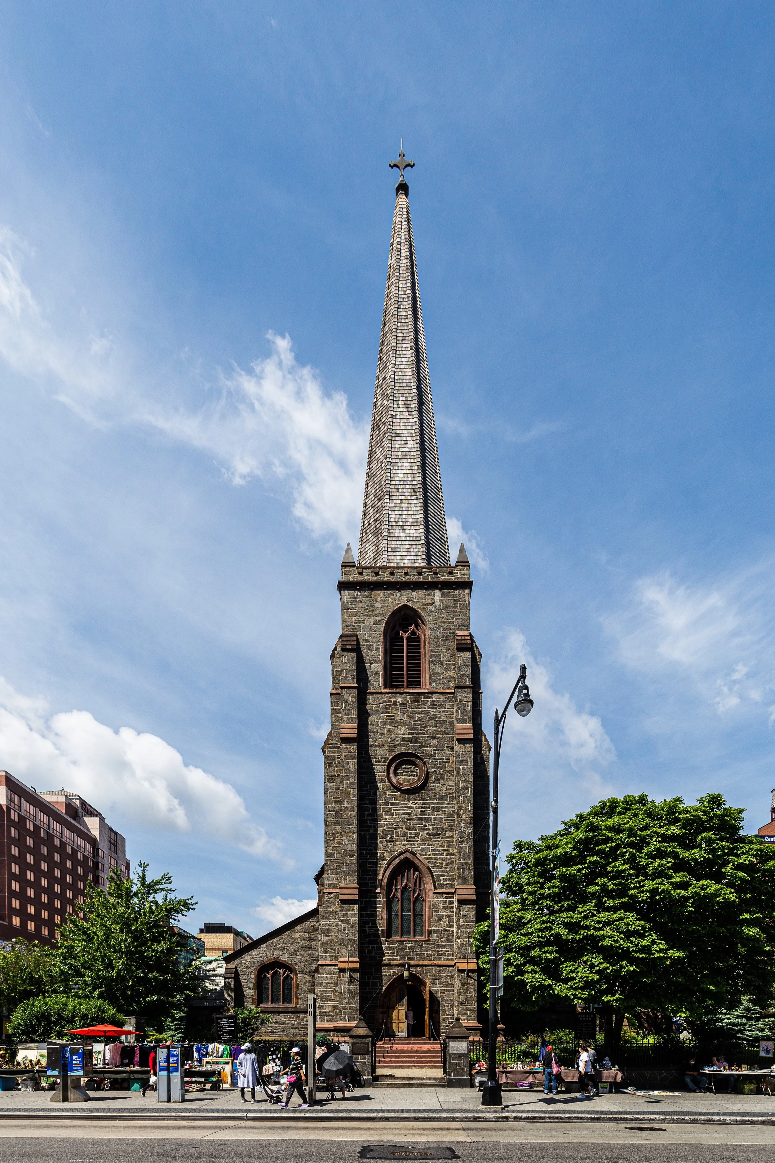 A tall stone church with a pointed steeple against a blue sky, with people walking in front and trees on the sides.
