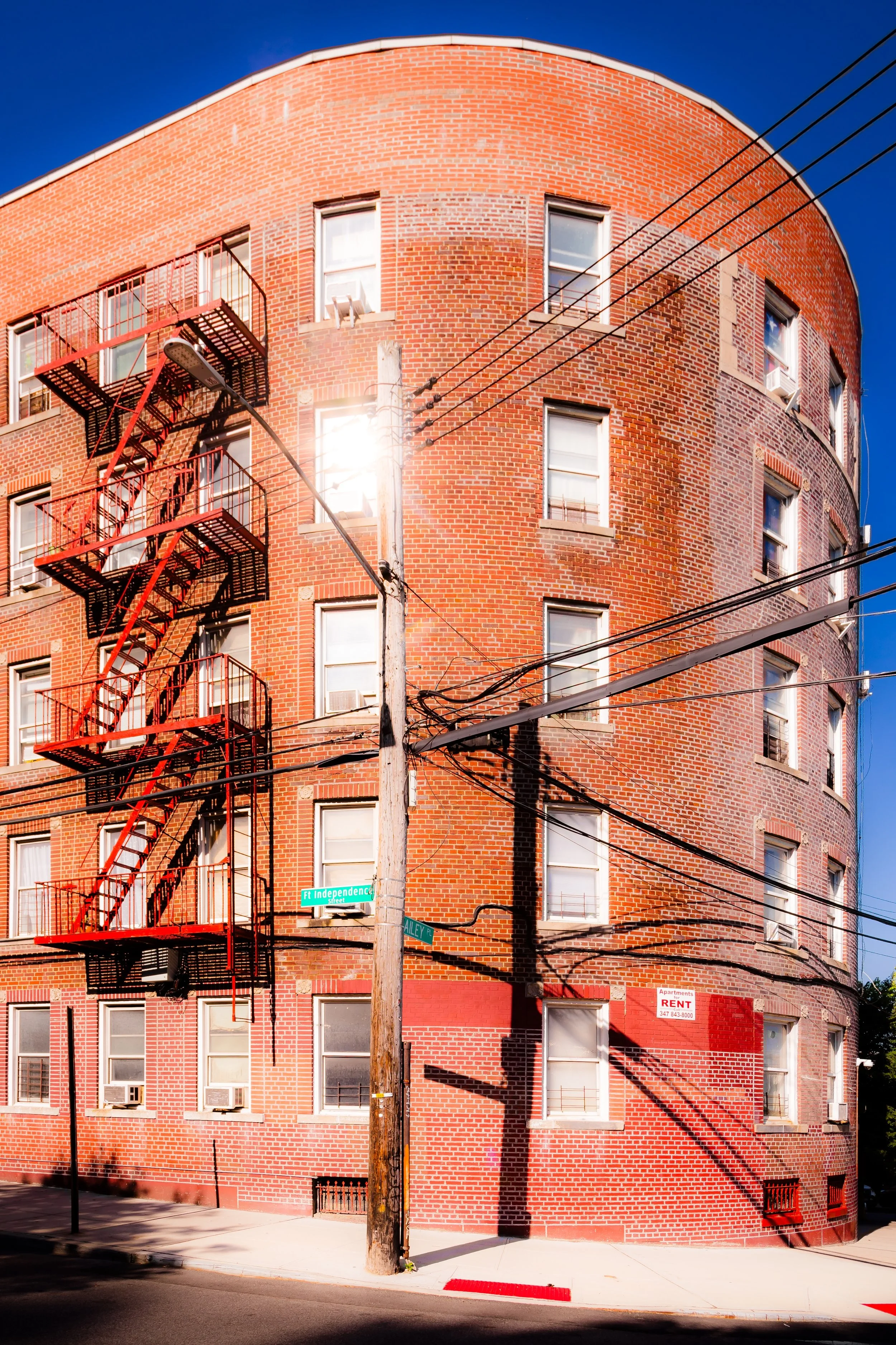 Red brick apartment building at street corner with fire escape, utility pole, and overhead wires in sunlight