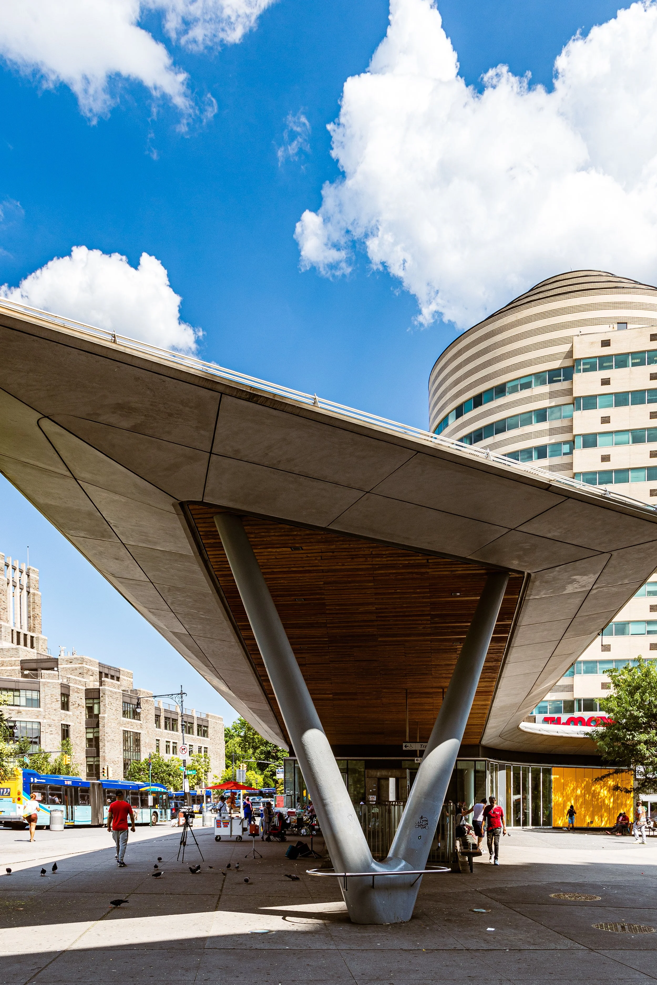 Modern urban scene with a large, curved building in the background, a shelter with angled supports in the foreground, blue sky with white clouds, and people walking along the street with trees and a bus.