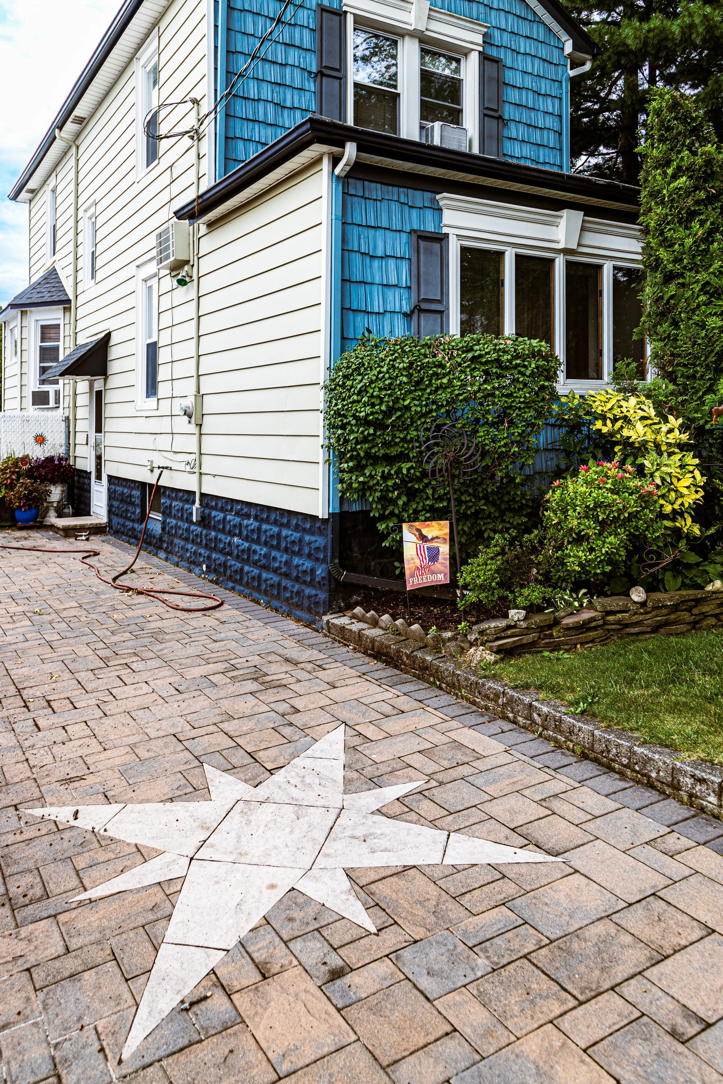 A house with a blue and white exterior, featuring multiple windows, some with black shutters, and a small front garden with greenery and a sign that reads "FREEDOM" with an image of an eagle and the American flag. The paved sidewalk in front has a white star design.
