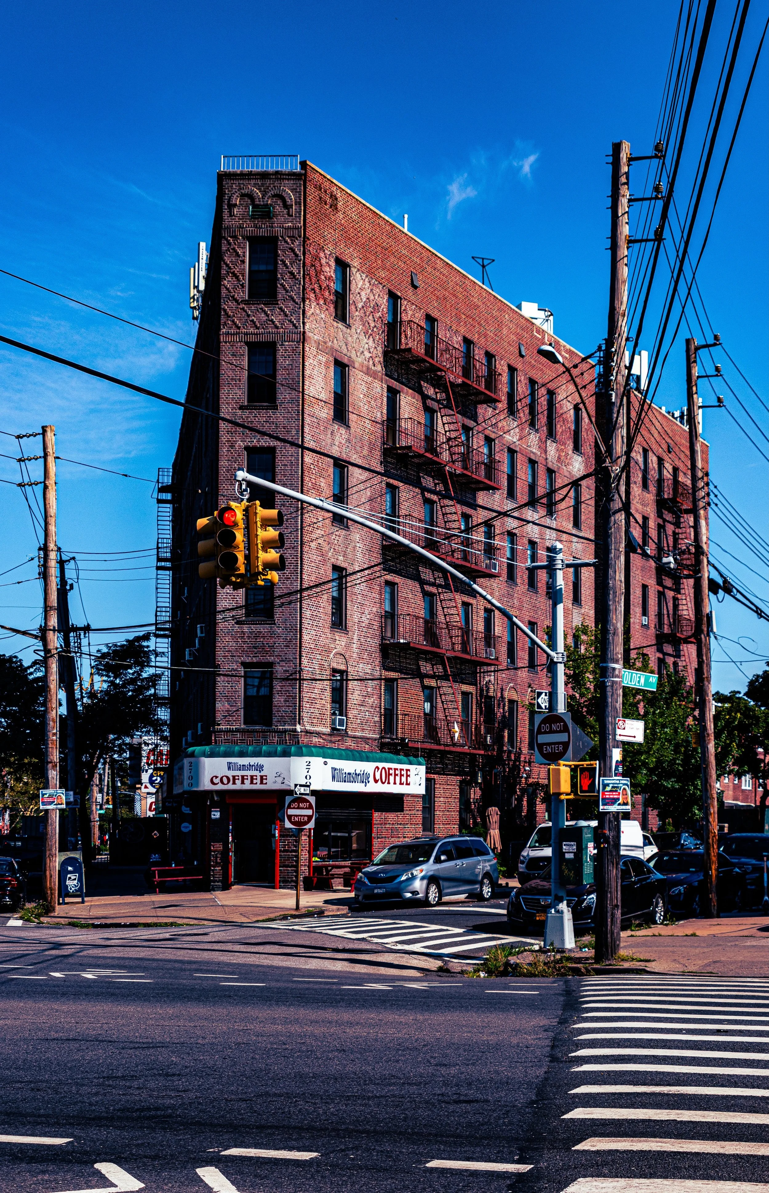 A tall red brick building with fire escapes, next to a street with parked cars and a crosswalk, under a clear blue sky.