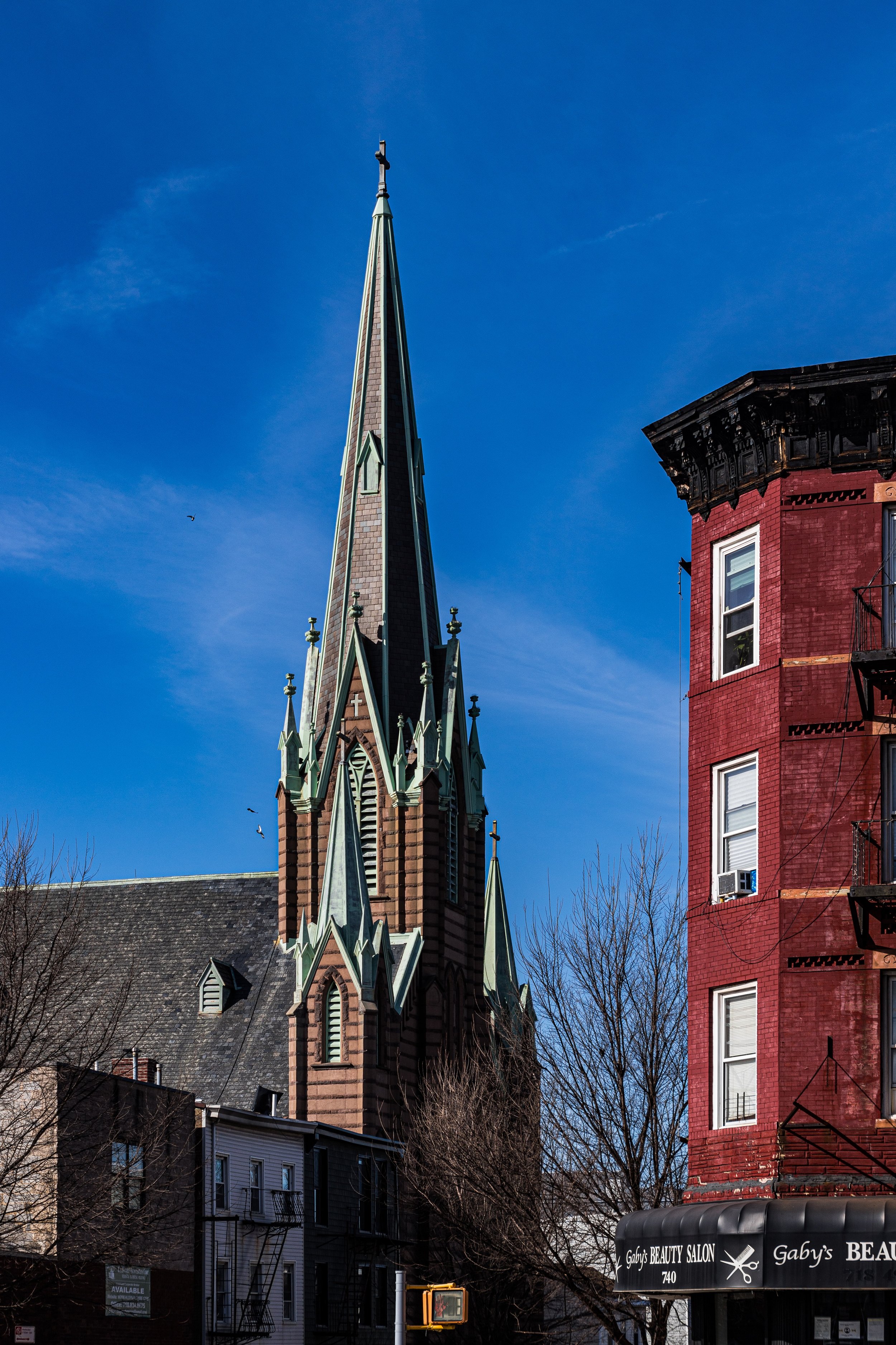 A tall Gothic-style church steeple with intricate designs, set against a bright blue sky, with a red brick building and leafless trees in the foreground.