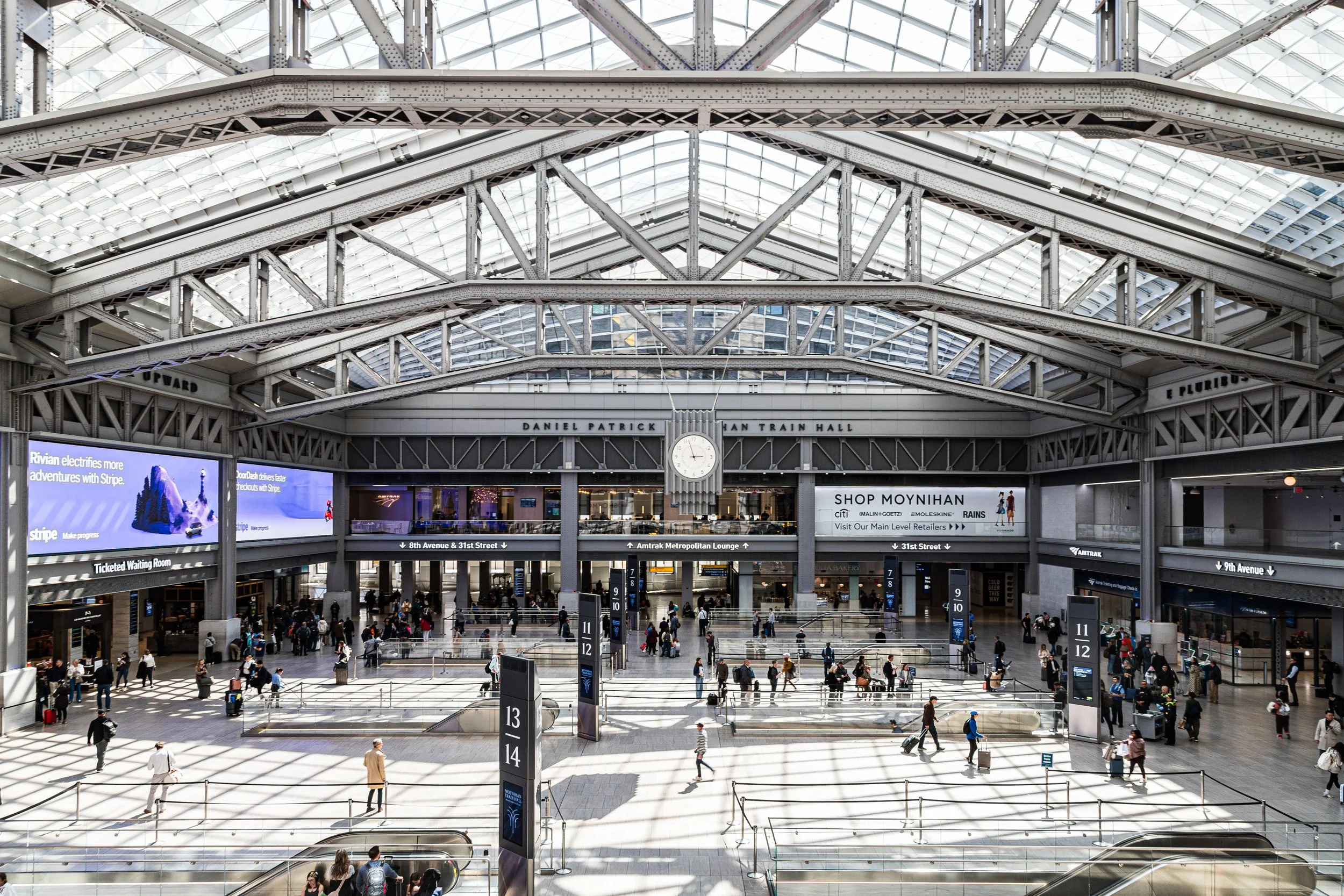 Interior of a large train station with high glass ceiling, crowds of travelers, and signs for platforms and services.