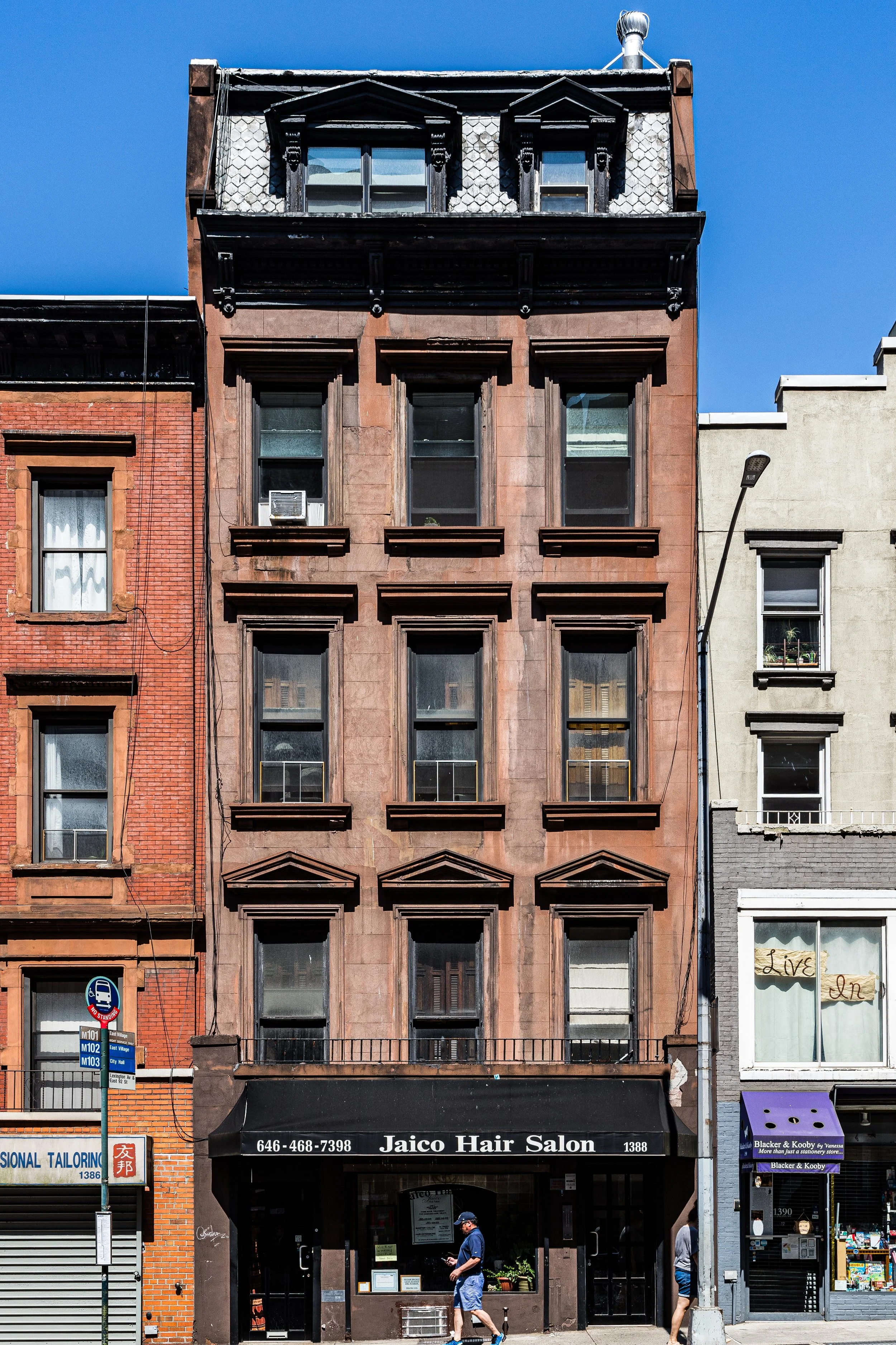 Tall brownstone building with multiple windows, some with air conditioning units, and a black awning that says Jaico Hair Salon at street level. A man is walking in front of the salon and a woman is walking past.