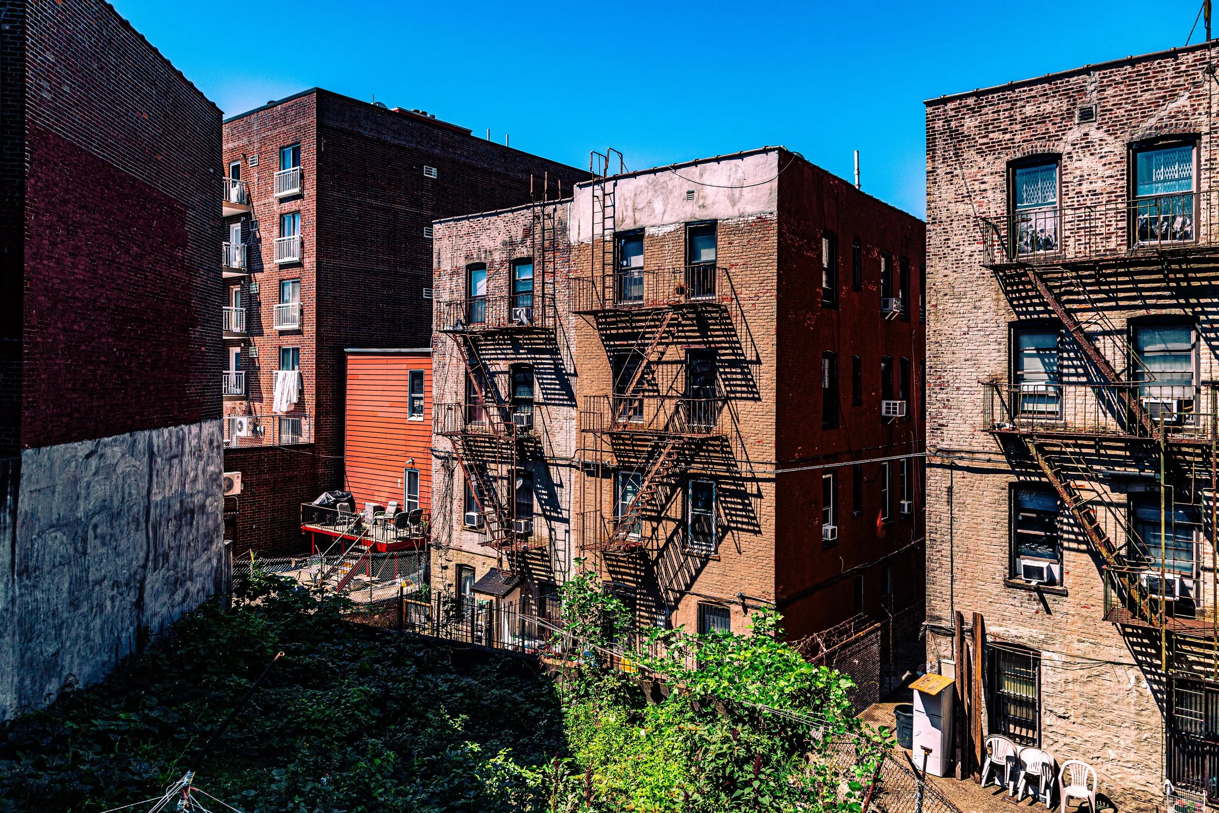 View of an urban courtyard surrounded by brick apartment buildings with fire escapes, windows, and balconies, with some trees and chairs in the foreground under a clear blue sky.