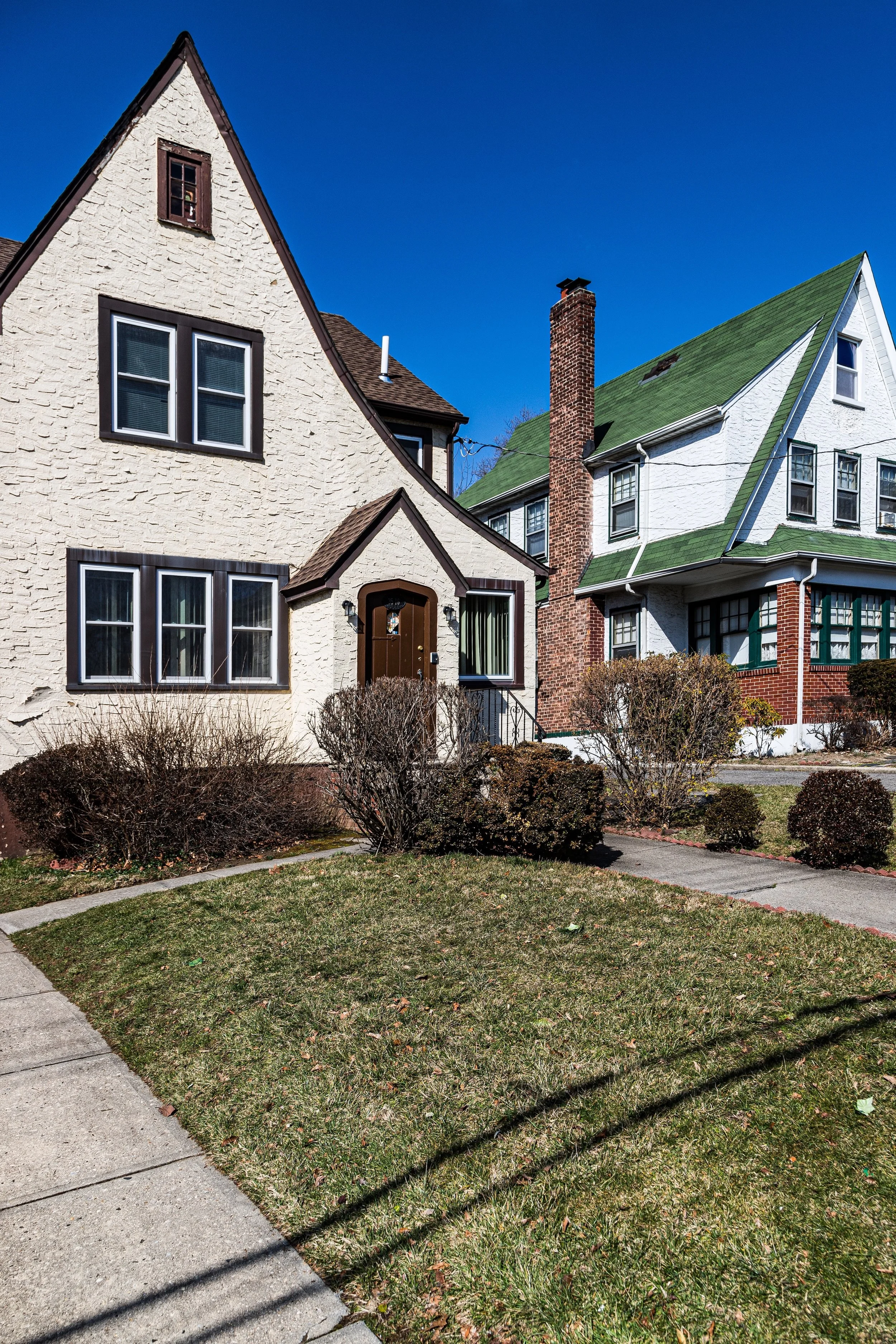 A residential street with two houses, one with a beige stucco exterior and a brown door, and a second with a white exterior and green roof. The front yard has grass and trimmed bushes, with sidewalk in the foreground and a clear blue sky overhead.
