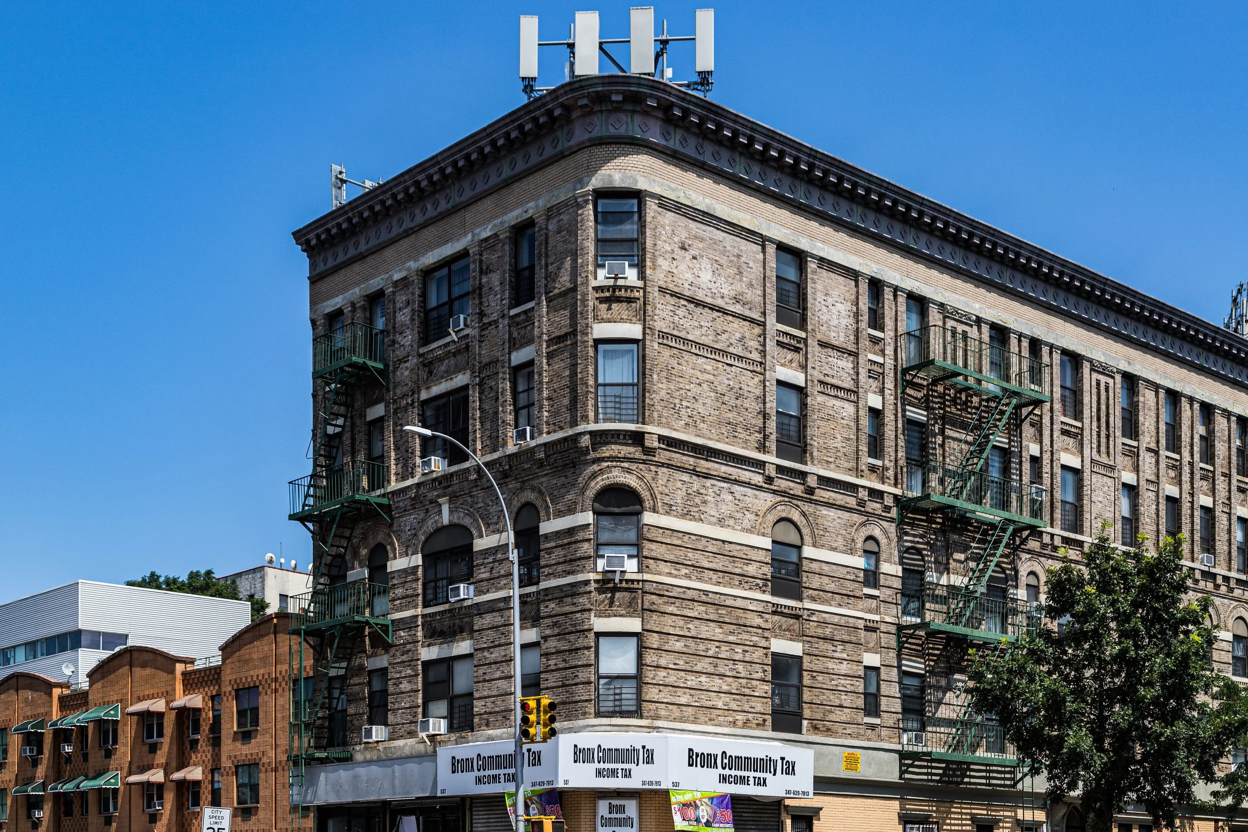 A multi-story brick building with fire escapes on the side, located at the corner of a city street, with a Bronx Community Tax sign at the lower level and a blue sky above.
