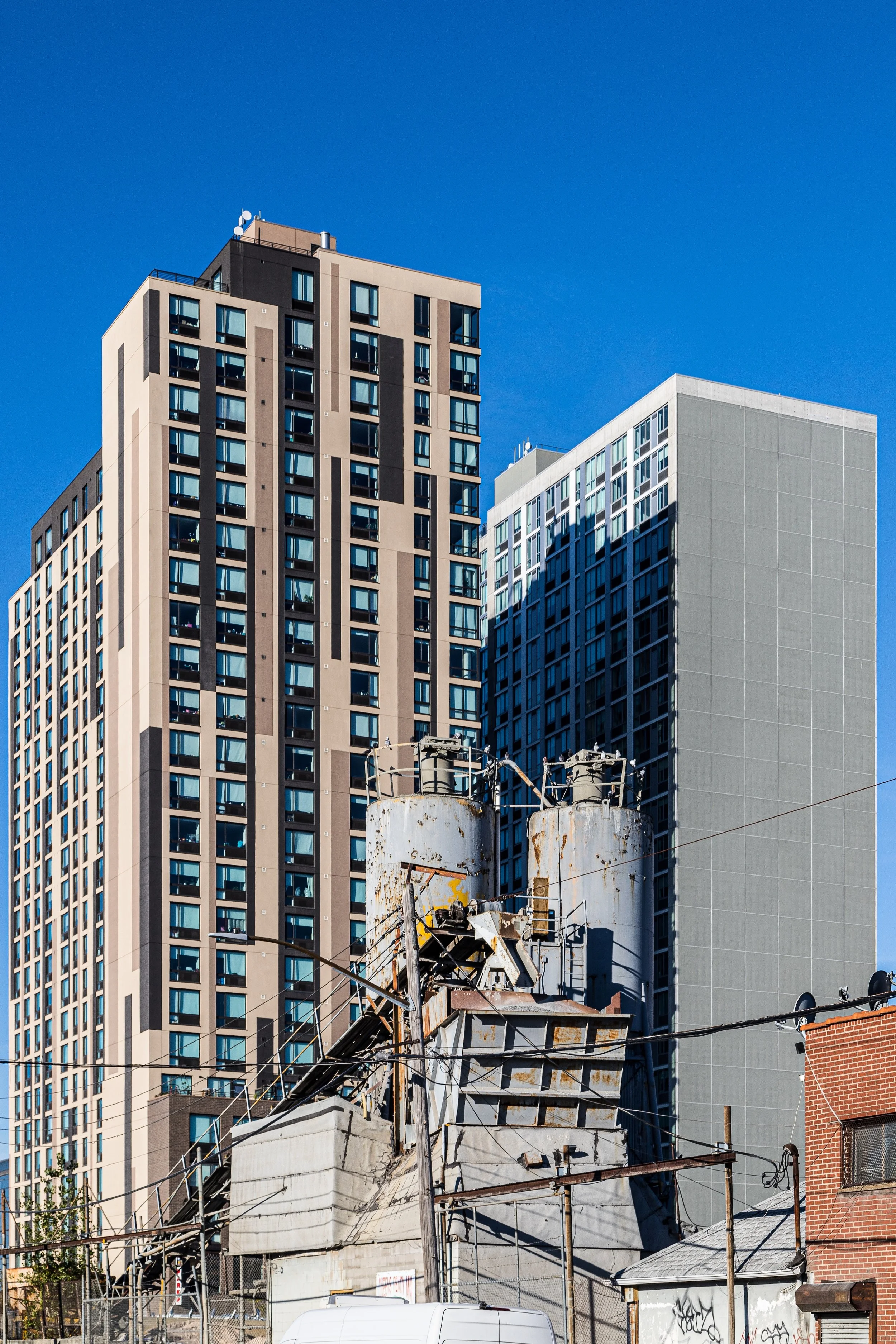 Old rusted industrial structures with two large tanks and electrical wires in front of modern high-rise buildings against a clear blue sky.