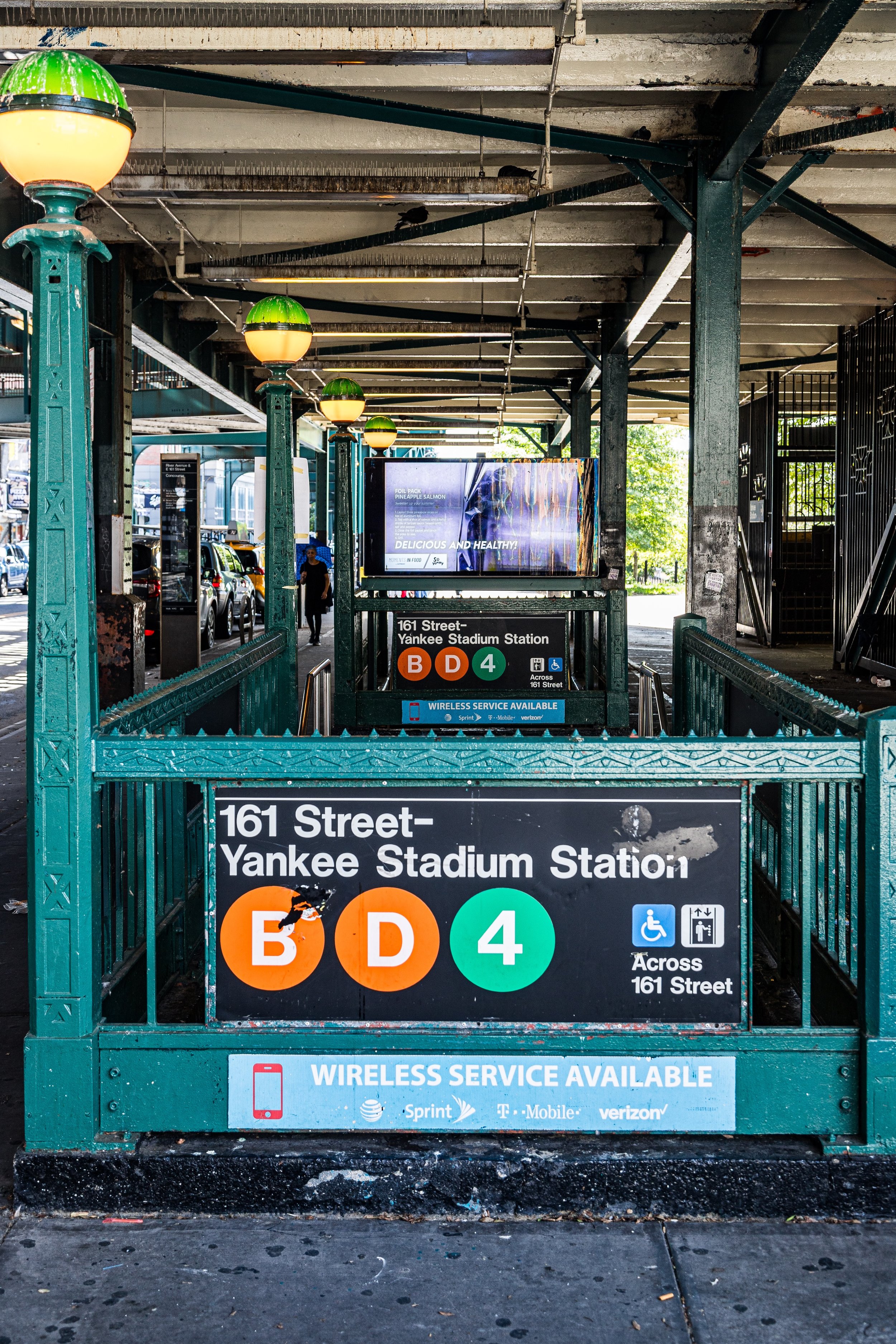 Entrance to the subway station at 161 Street-Yankee Stadium, showing signs for B, D, and 4 subway lines, with an advertisement board and a blue information sign.