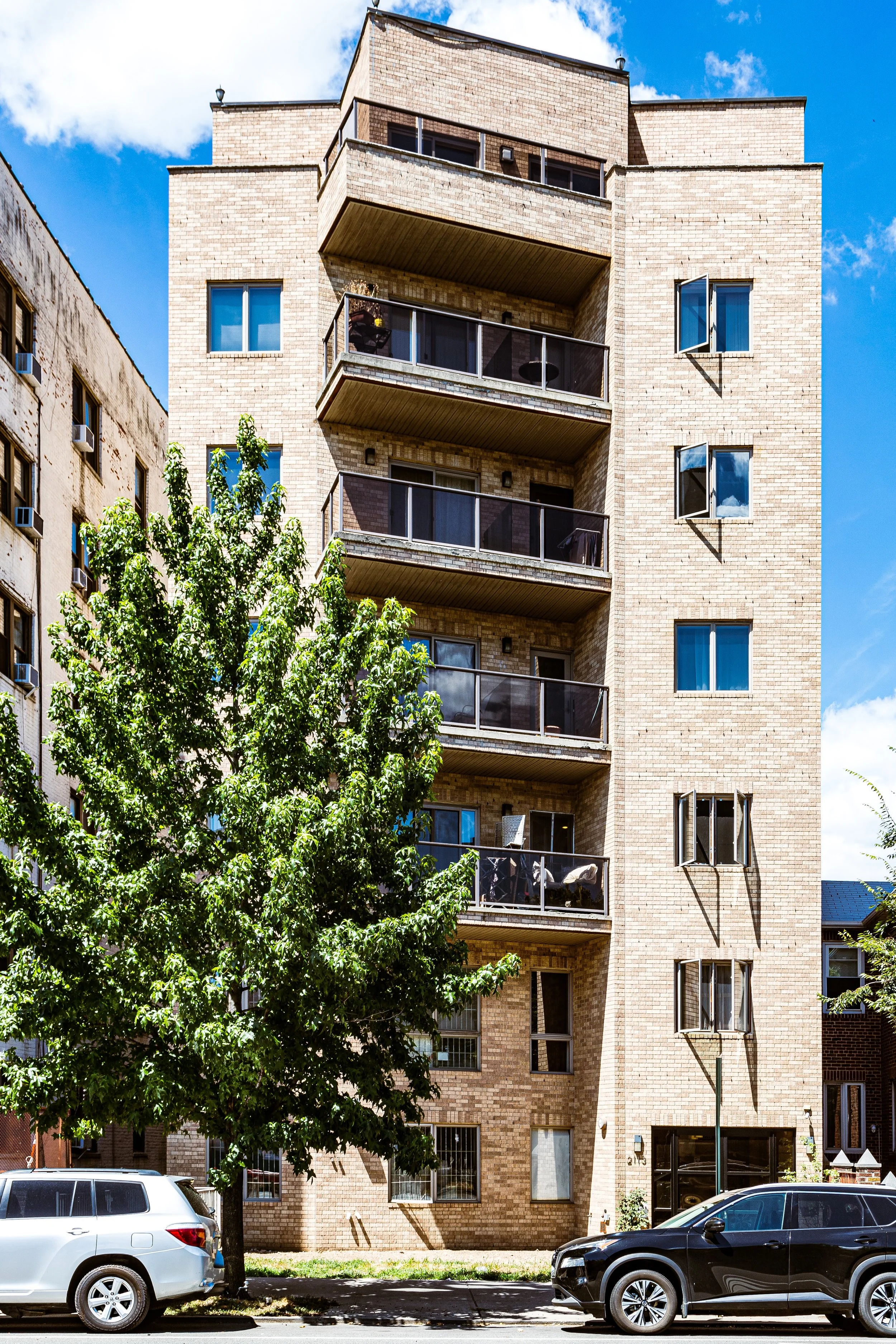 A multi-story brick apartment building with balconies on each floor. A green tree is in front, partially obscuring the building. Two cars, a white SUV and a black sedan, are parked on the street.