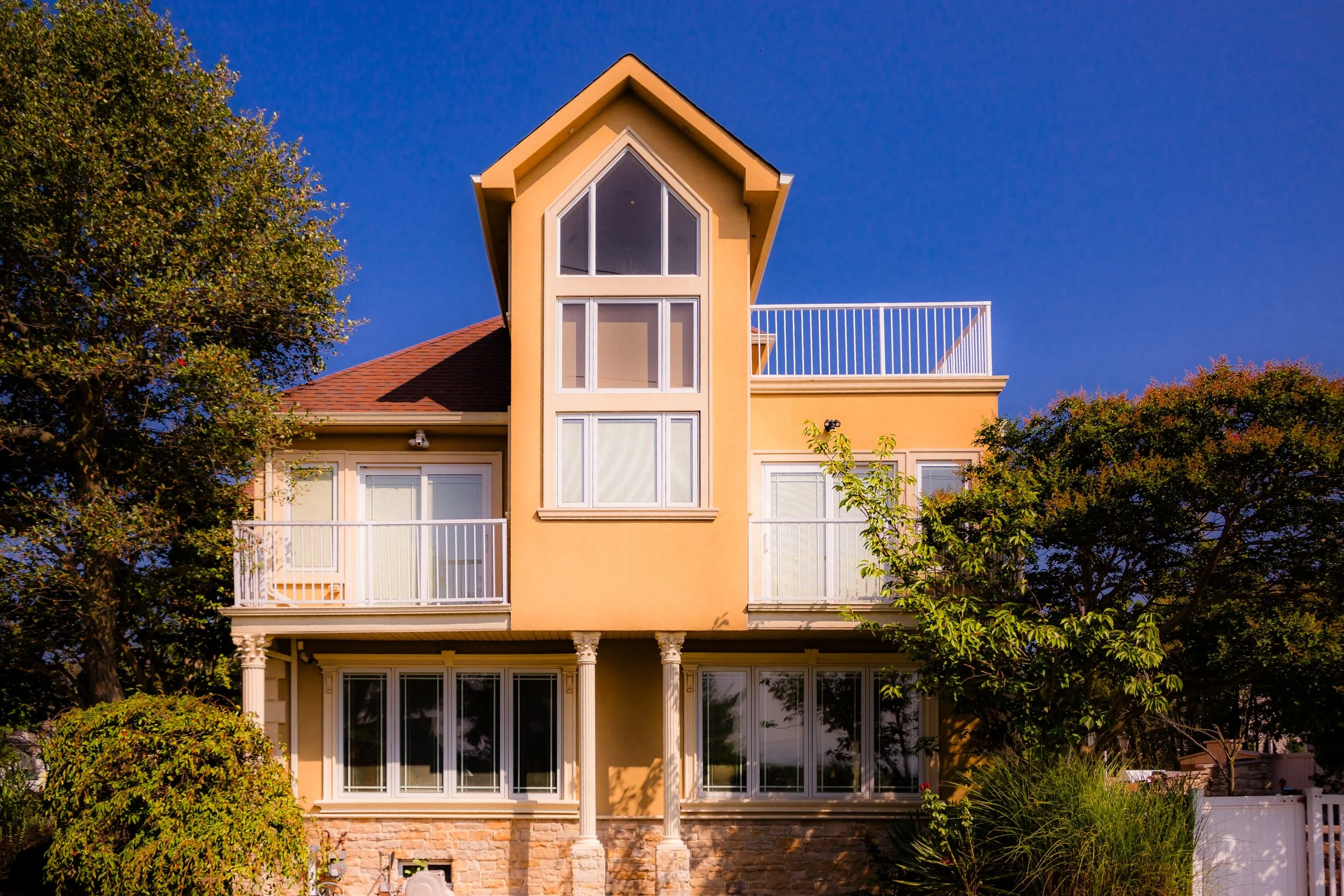 A multi-story house painted in beige with a distinctive tall, peaked tower featuring large windows. The house has balconies with white railings, a stone foundation, and is surrounded by green trees and bushes against a clear blue sky.