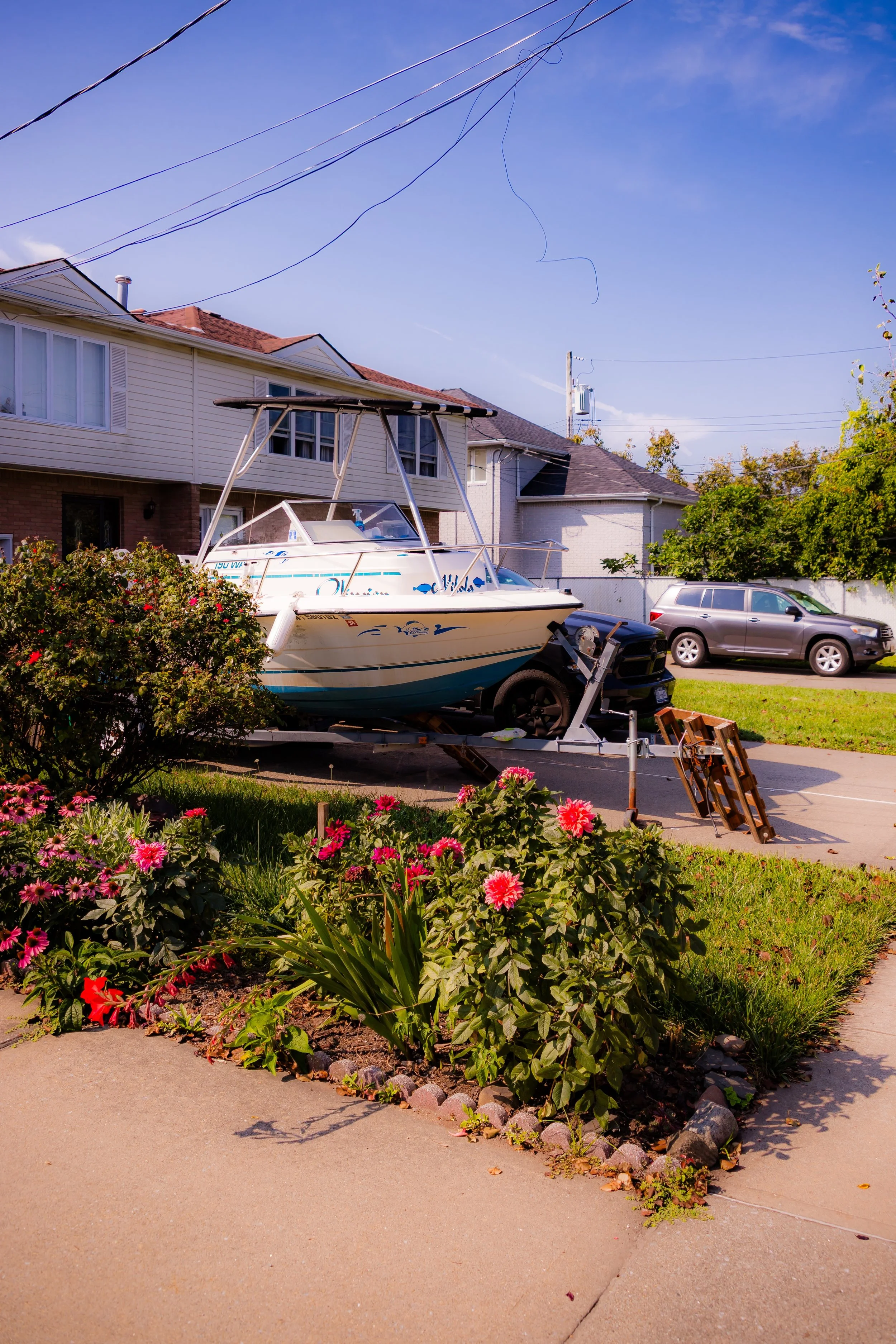 A boat on a trailer parked in a residential driveway with flowers and bushes in the foreground and houses and cars in the background.