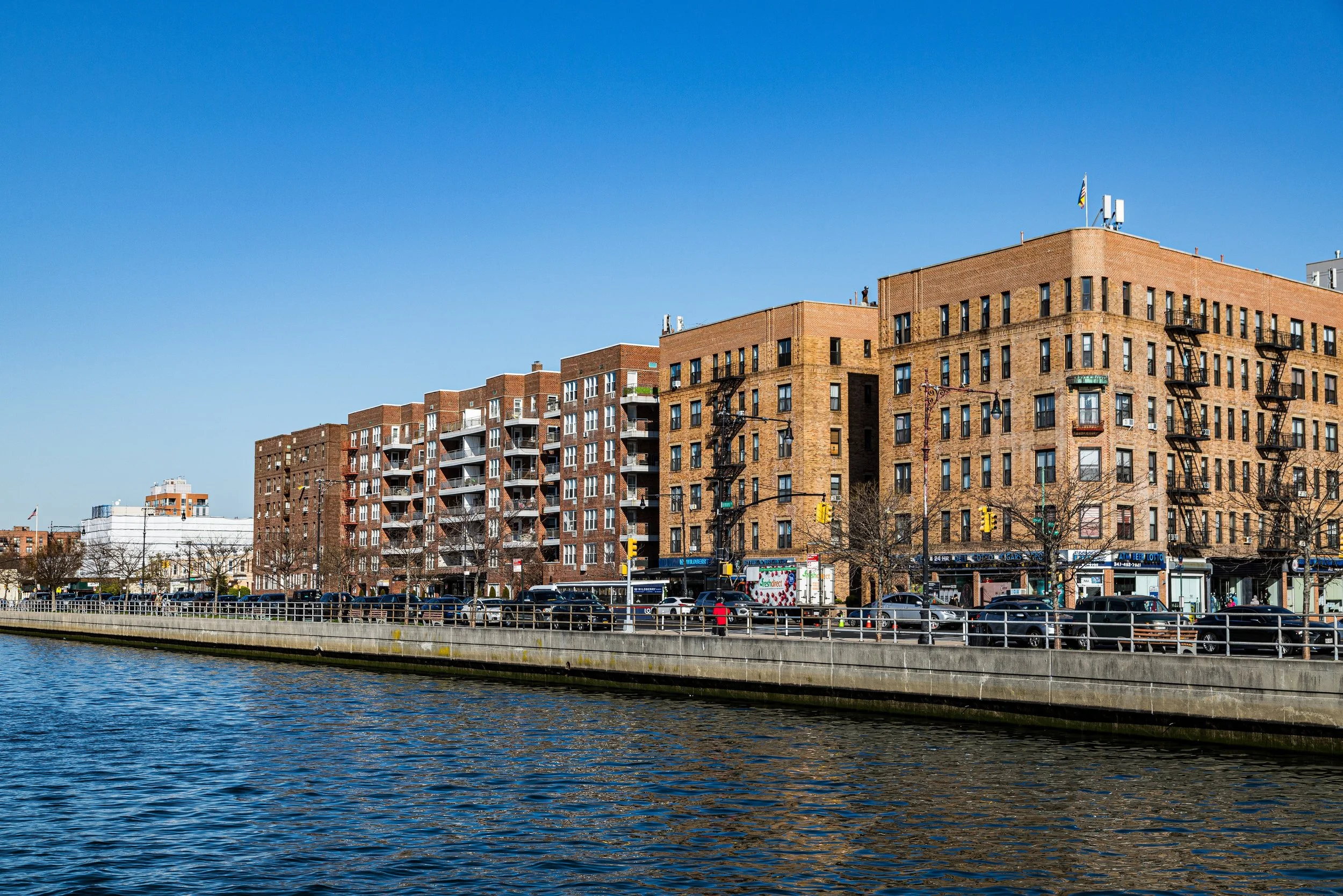 View of a city street with brick apartment buildings along a waterway under a clear blue sky.