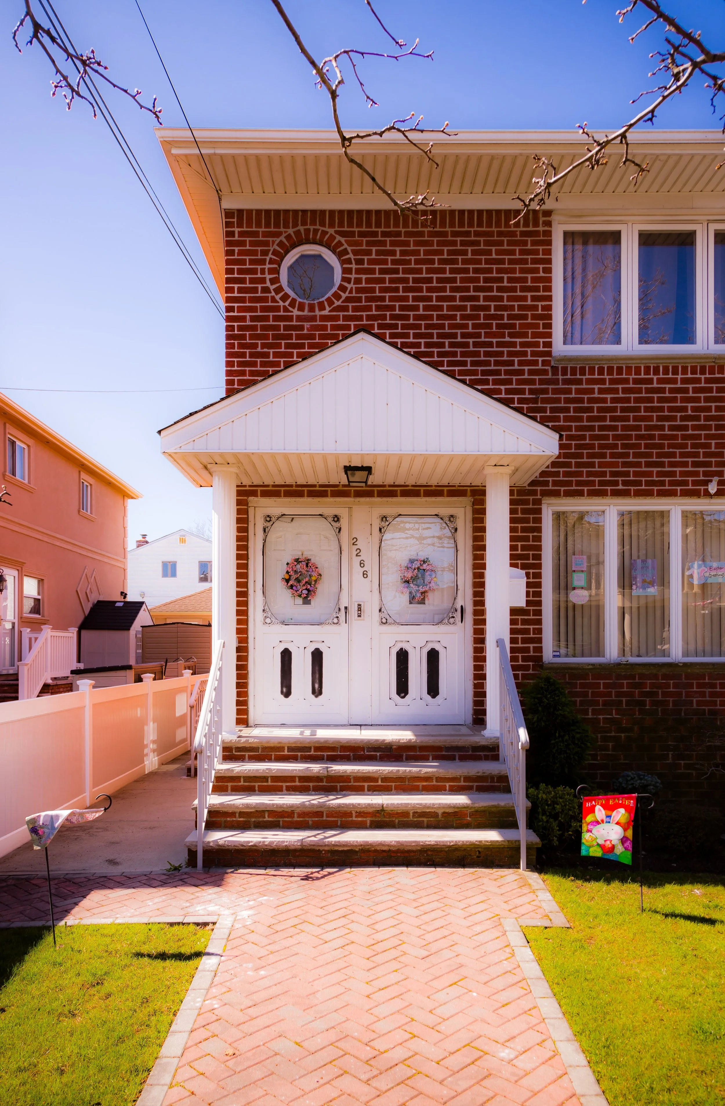 A brick house with white double front doors, decorated with Easter wreaths, and a small front yard with a brick walkway, Easter sign, and patches of grass.