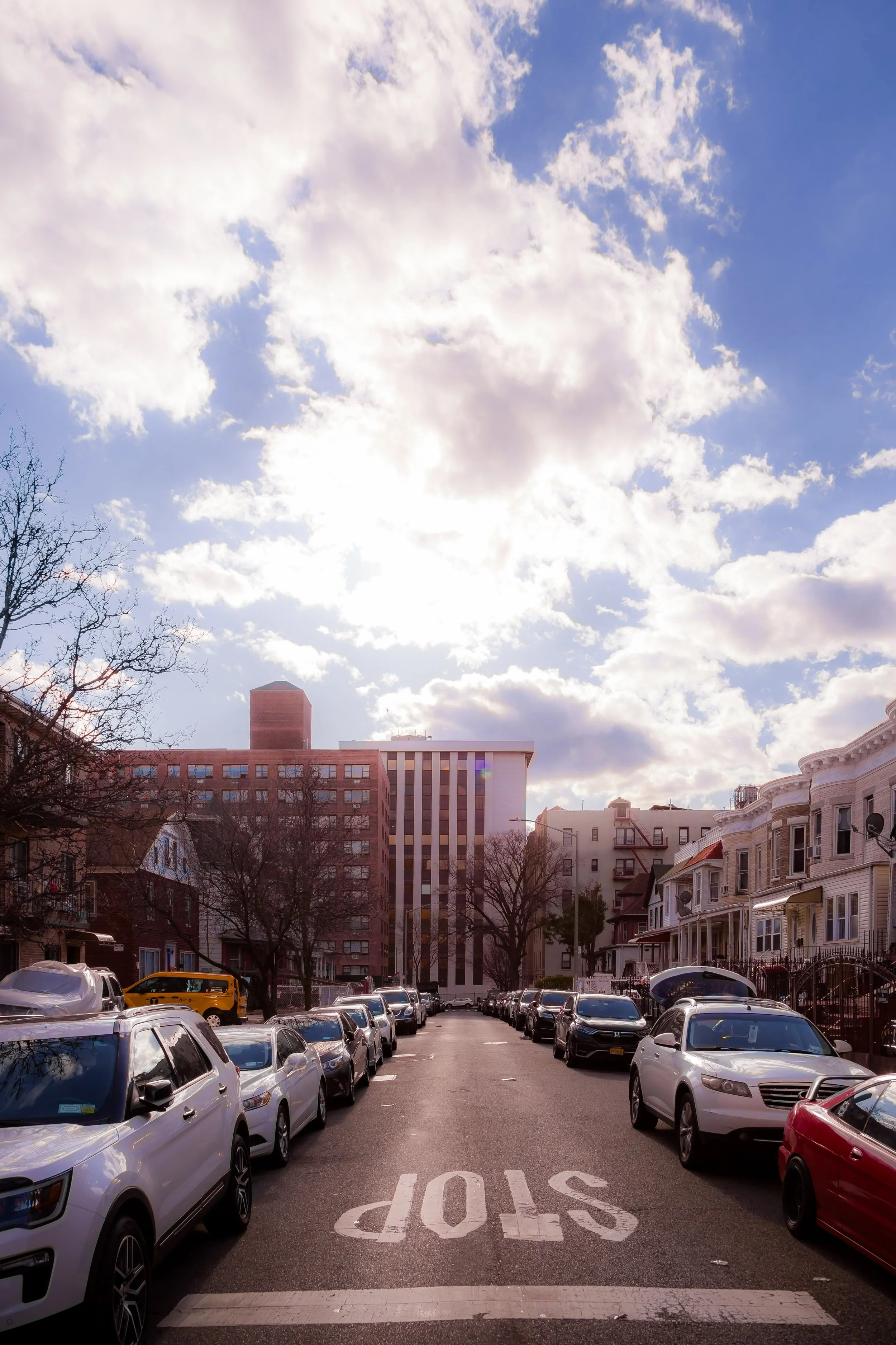 A city street with cars parked along both sides, residential houses on the right, and taller buildings in the background under a partly cloudy sky with the sun peeking through.