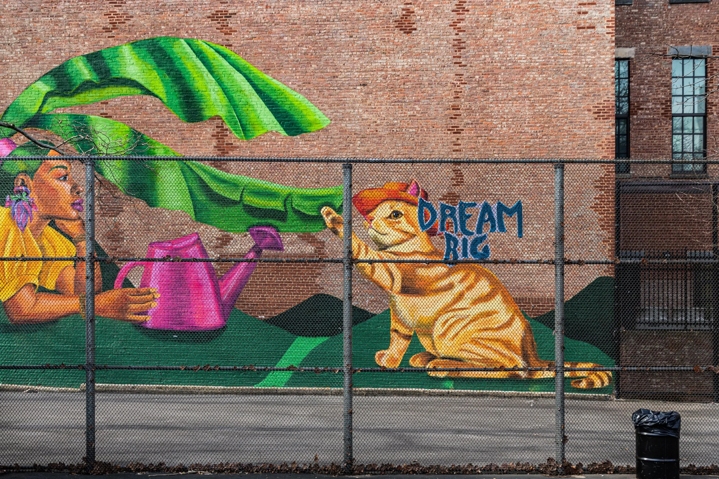 Colorful mural on a brick wall depicting a woman watering a watermelon and a tiger with the text 'Dream Big' beside it, behind a chain-link fence.