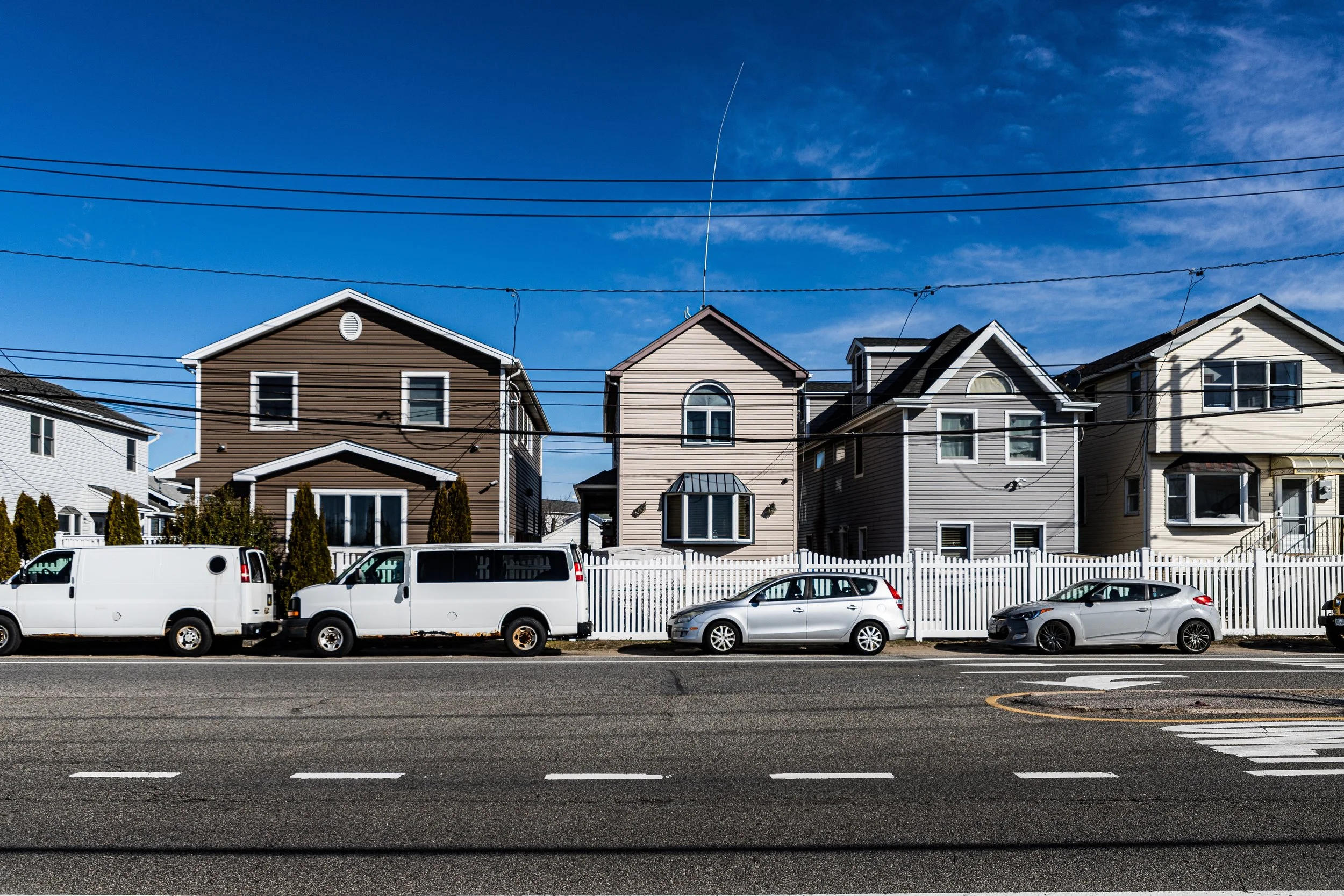 A row of four residential houses with a fence in front, parked cars on the street, and a clear blue sky overhead.
