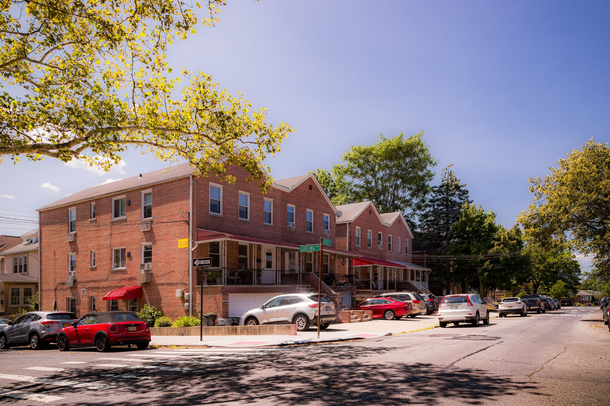 A street scene with brick townhouses, parked cars, trees, and a blue sky.