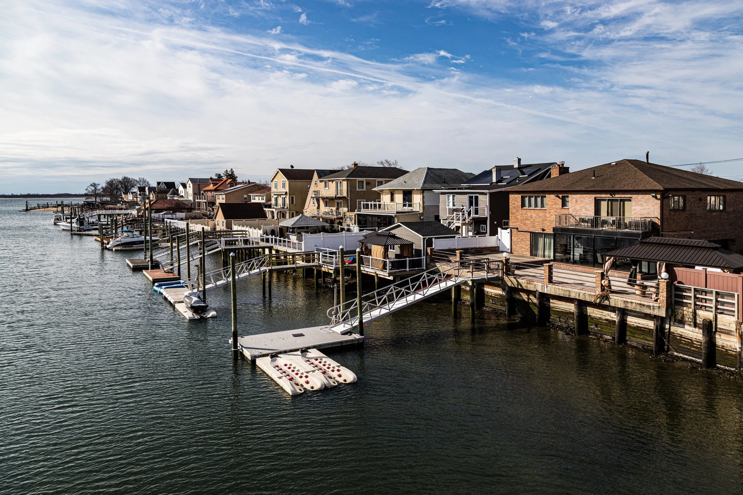 A row of houses along a waterway with docks and boats, under a partly cloudy sky.