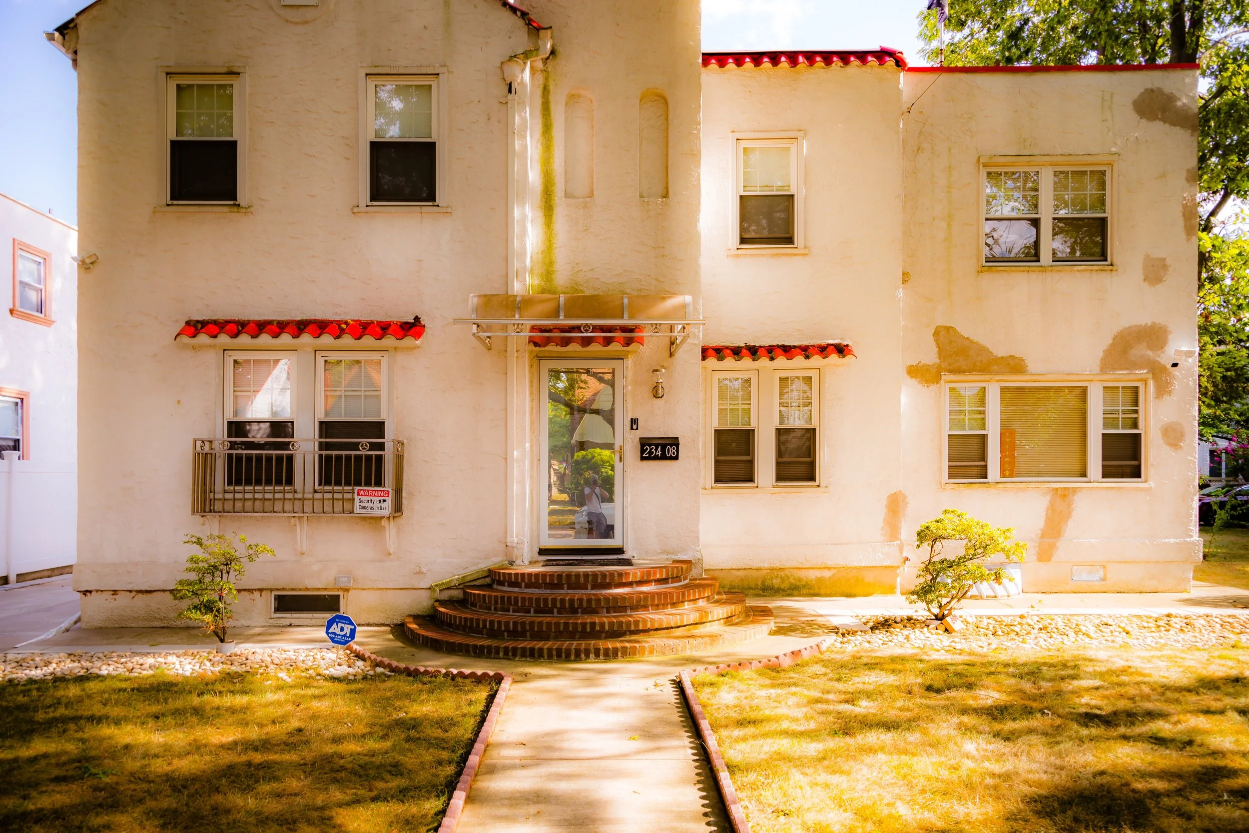 Front view of a two-story house with a white stucco exterior, red-tiled awnings above the windows, and a brick staircase leading to a glass front door. The house has multiple windows and a small garden with shrubs and a sidewalk.